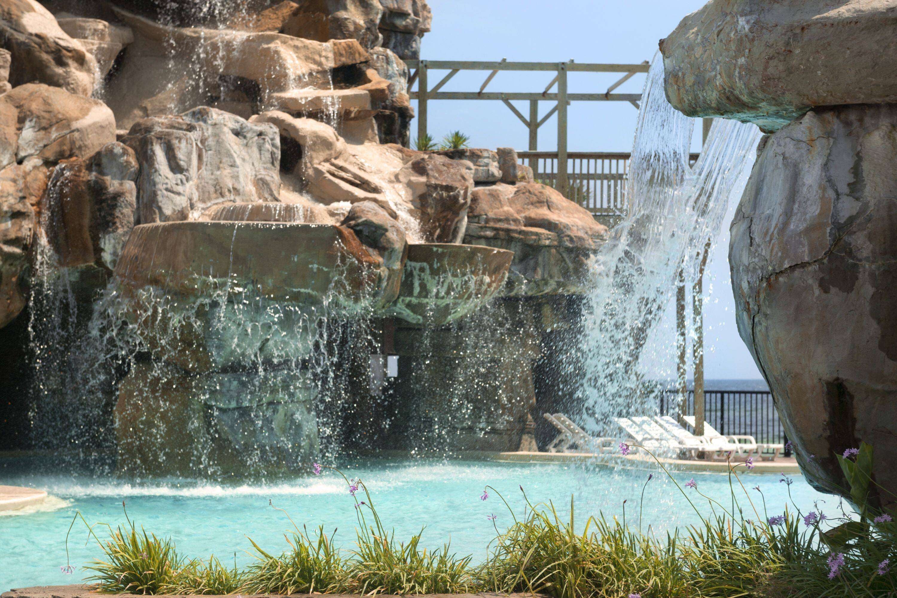 A pool with a waterfall feature surrounded by large rocks and a wooden deck with lounge chairs.