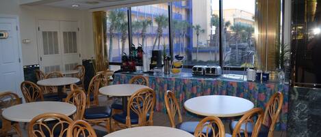A restaurant with round tables and wicker chairs, a counter with coffee machines, and a view of palm trees and buildings through large windows.