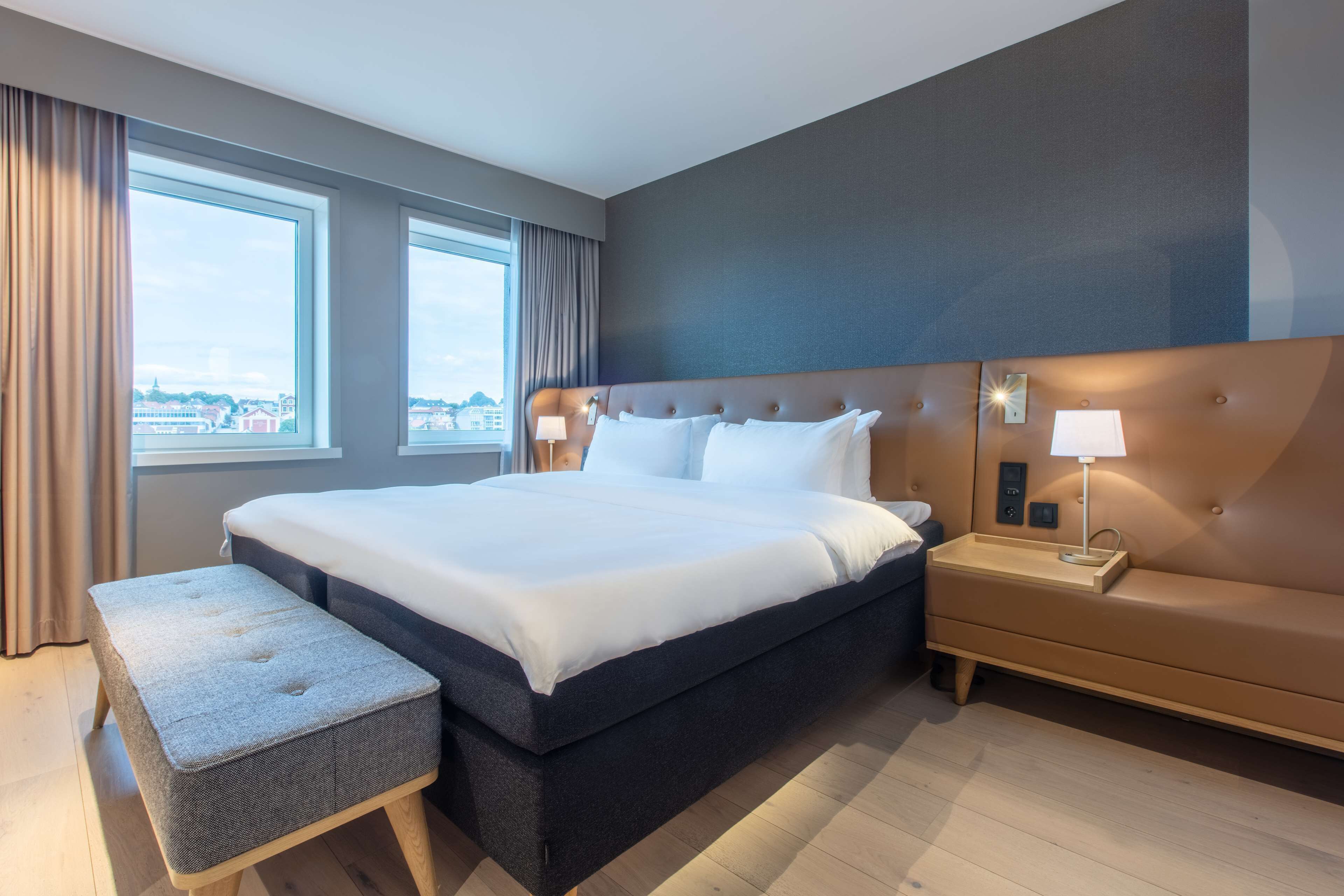 Hotelkamer met een groot bed, nachtkastje met lamp, een bankje en uitzicht op de stad door grote ramen.