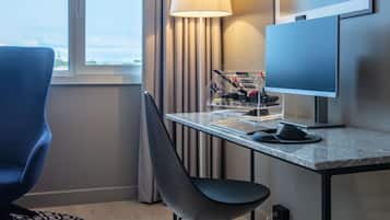 Habitación de hotel con escritorio, silla, lámpara, computadora y vistas de la ciudad a través de la ventana.