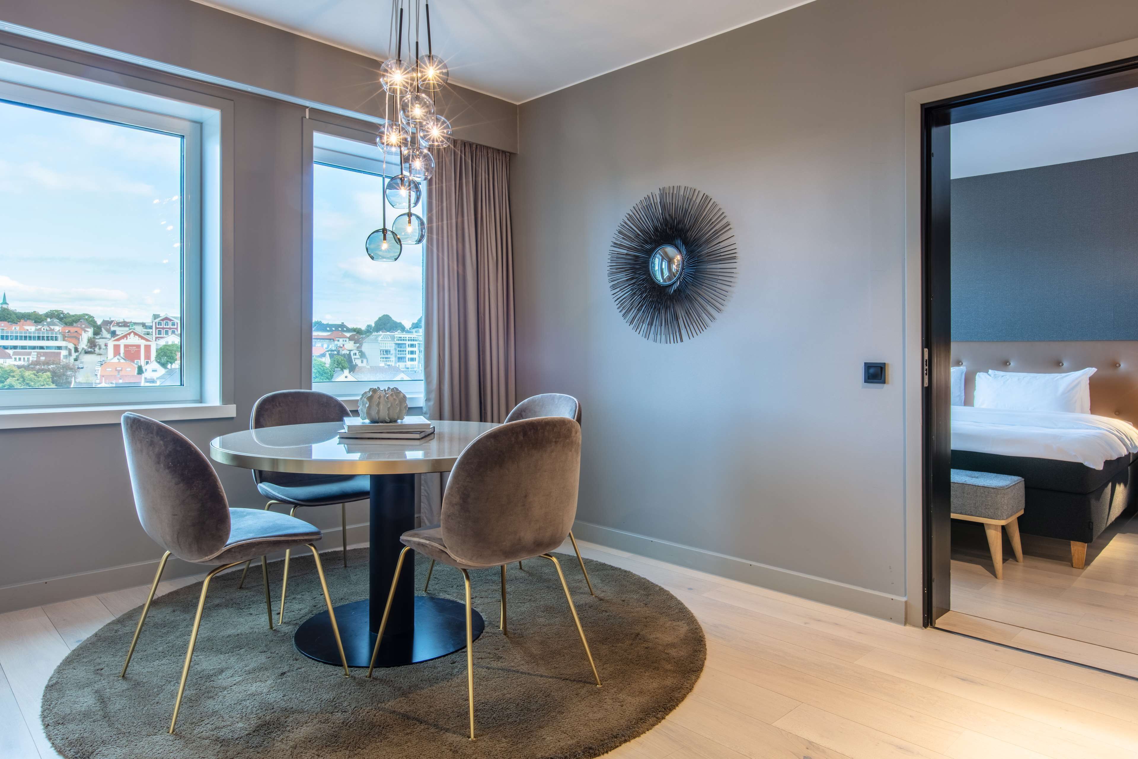 Una habitación de hotel moderna con una mesa de comedor redonda, dos sillas, un candelabro, un ventanal con vistas a la ciudad, un adorno circular en la pared y una zona de dormitorio separada con una cama y un banco.