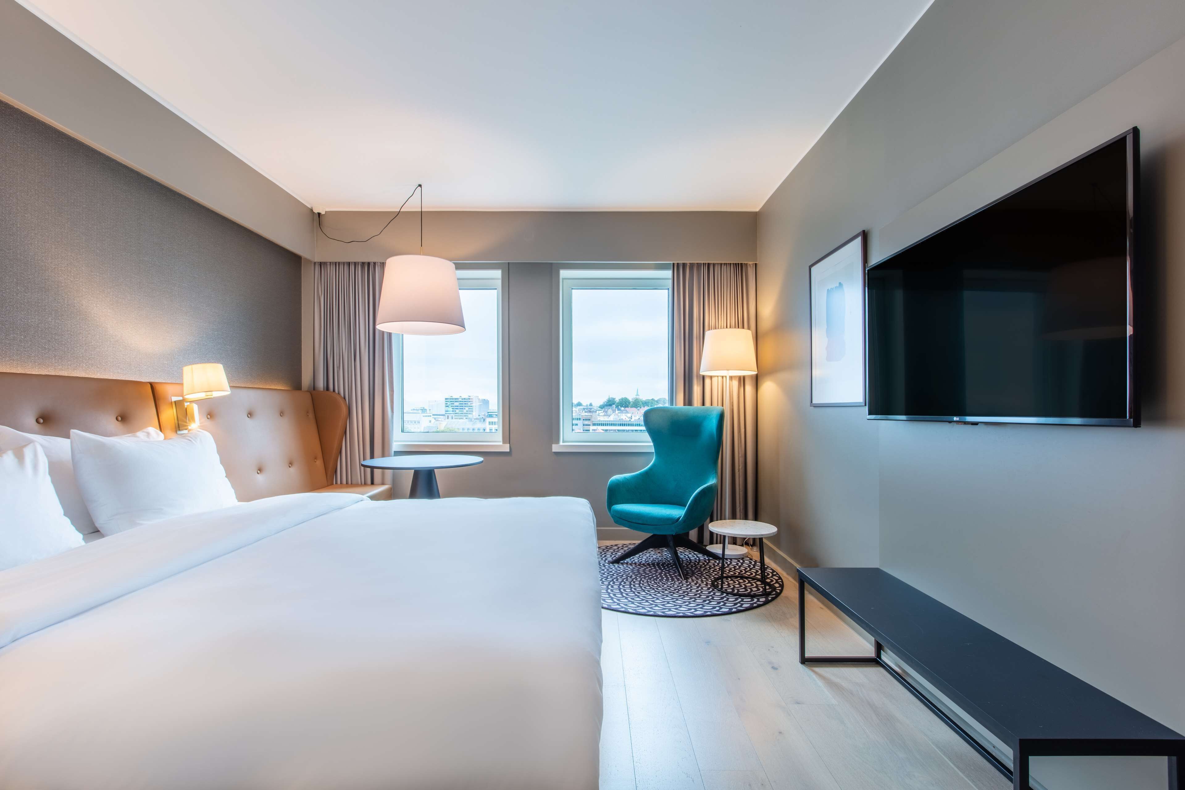 A hotel room with a large bed, a flat-screen TV, a teal armchair, and two windows with curtains.