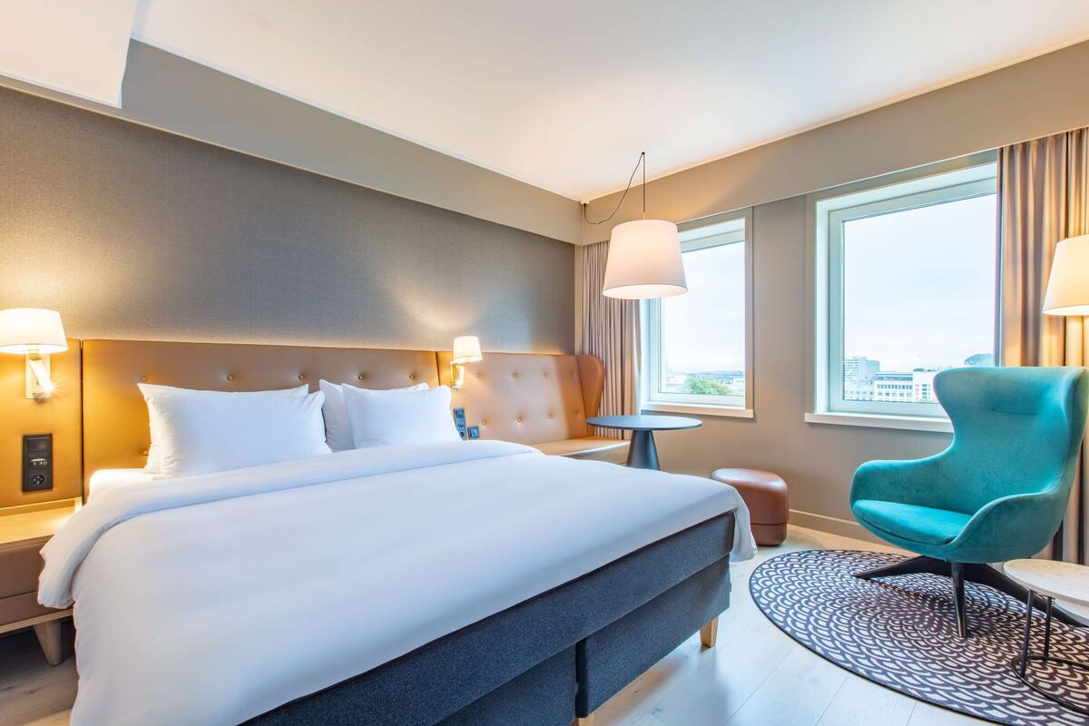Hotellihuone, jossa on suuri sänky, yöpöydällä olevat lamput, pyöreä pöytä, turkoosinsininen nojatuoli ja ikkunasta avautuva näkymä kaupunkiin.