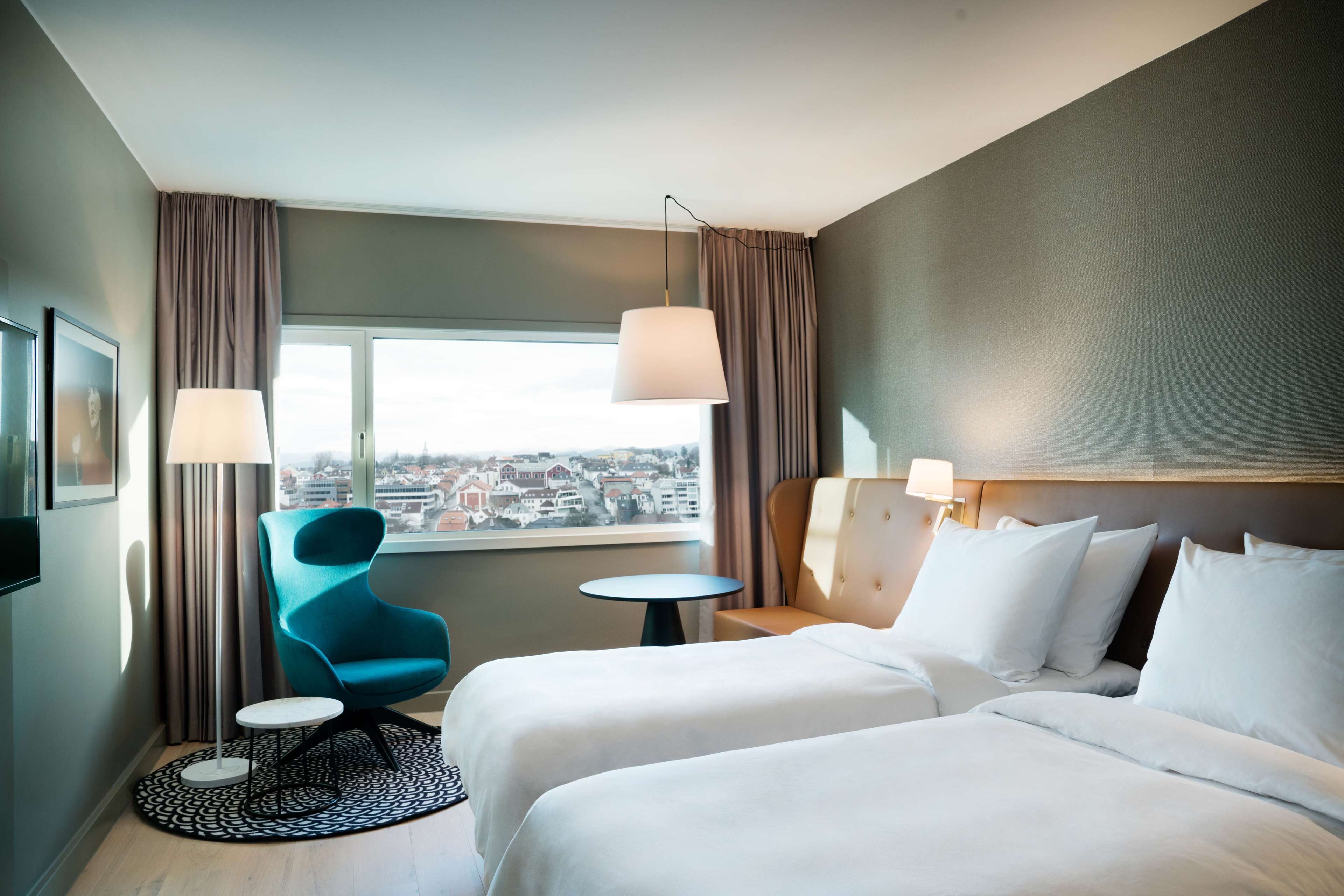 Habitación de hotel con dos camas, vista a la ciudad, una silla moderna y una mesita.