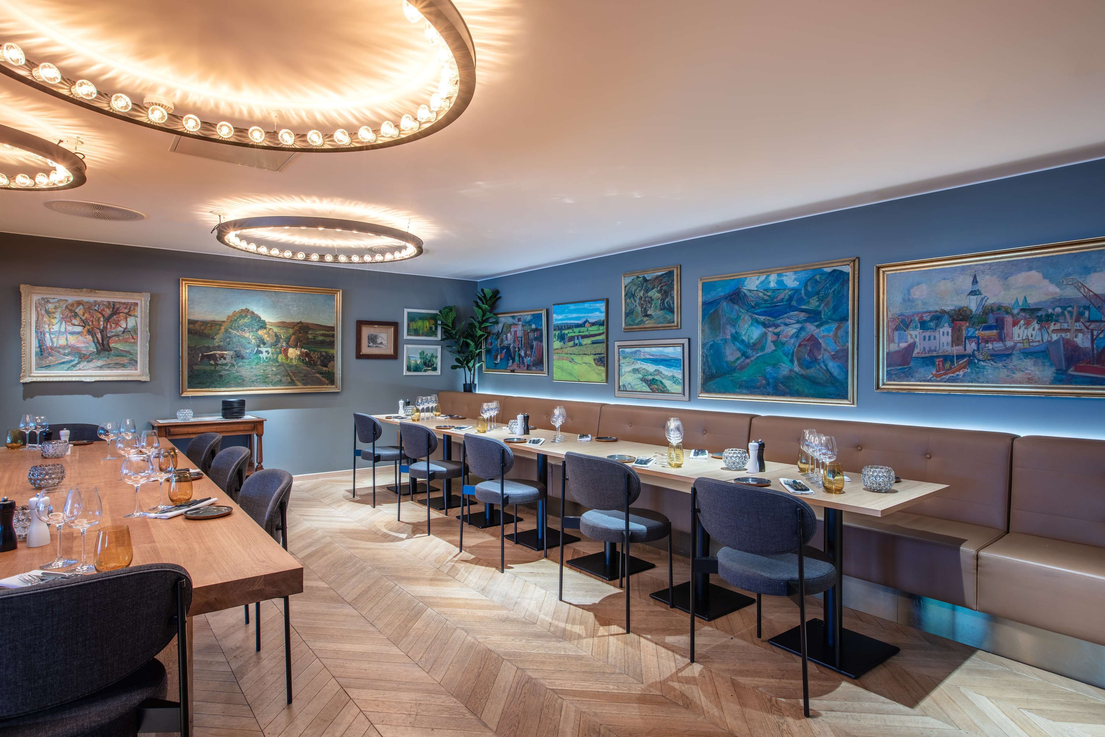 A modern dining area with a long wooden table, multiple chairs, and a bench. The room features a herringbone floor pattern and is decorated with framed artworks on the walls.