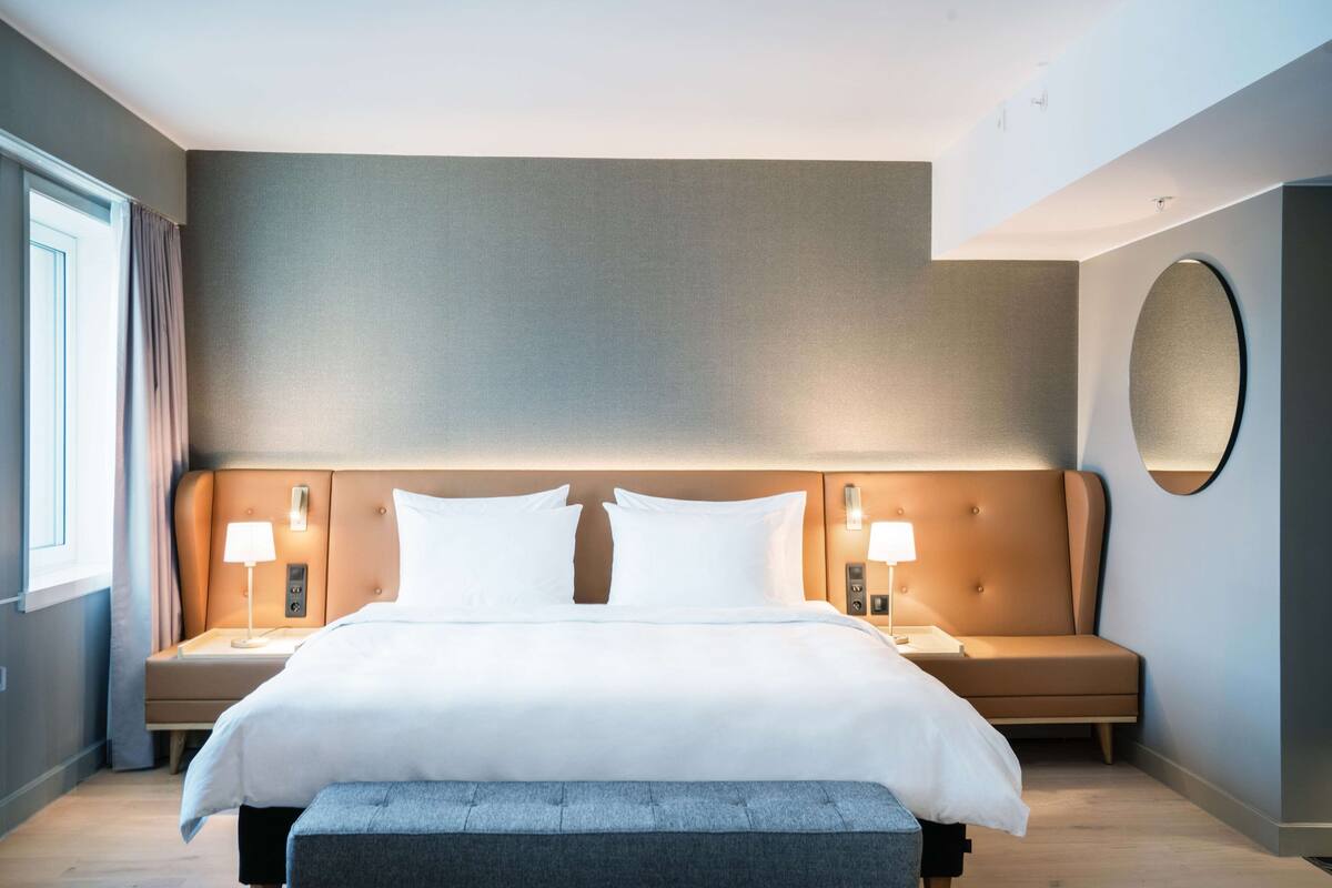Moderni hotellihuone, jossa on suuri sänky, kaksi yöpöytää valaisimineen, pyöreä peili ja penkki.