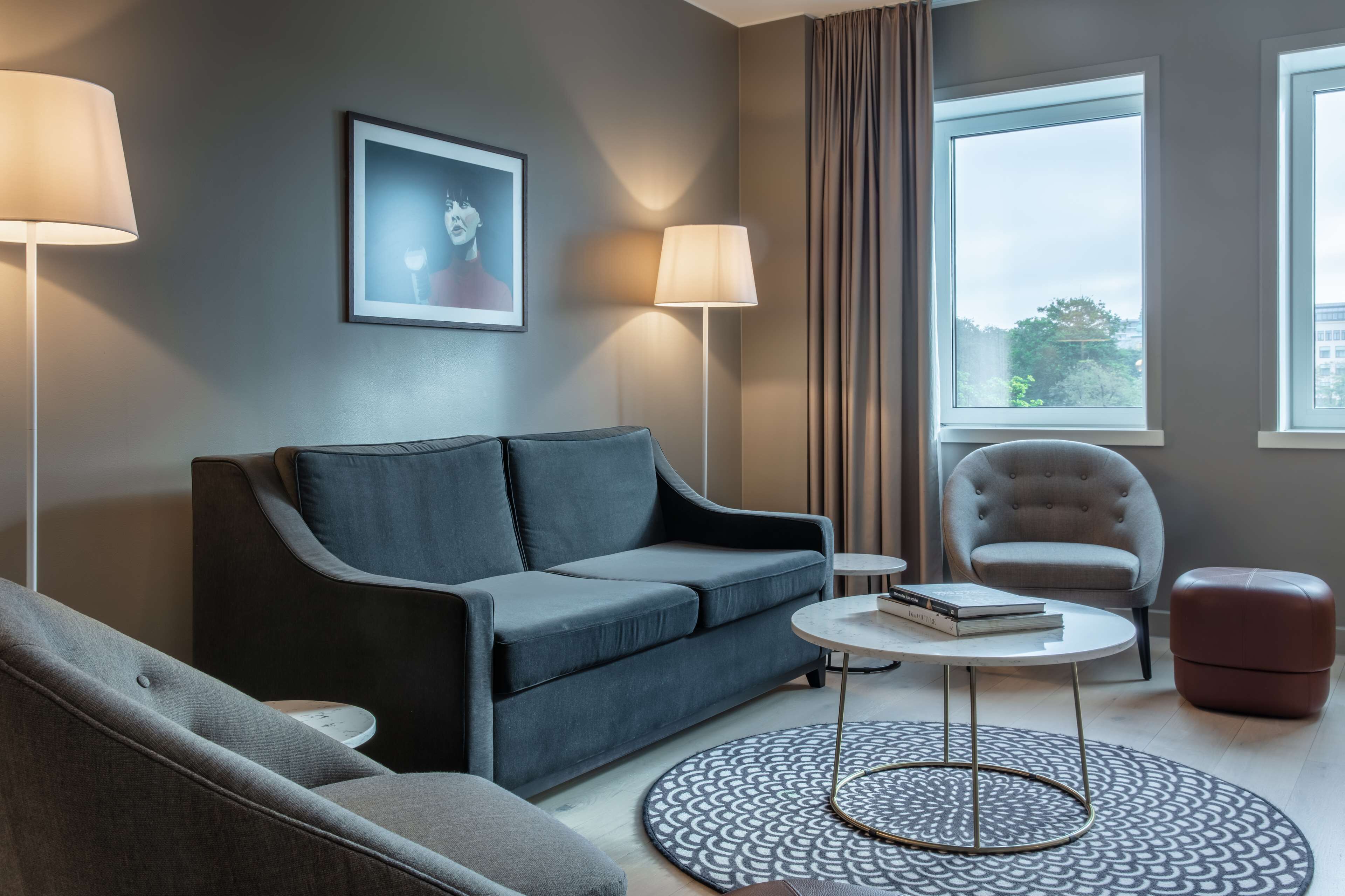A modern living room with a grey sofa, a round coffee table, a side table, a chair, and a window with curtains.