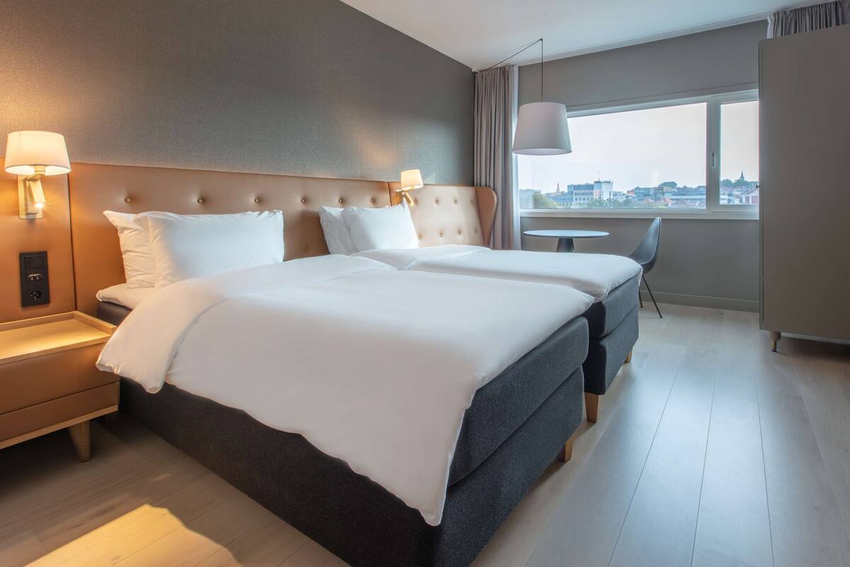 Hotellihuone, jossa on suuri sänky, yöpöydät, pieni pöytä ja ikkunasta avautuva näkymä kaupunkiin.