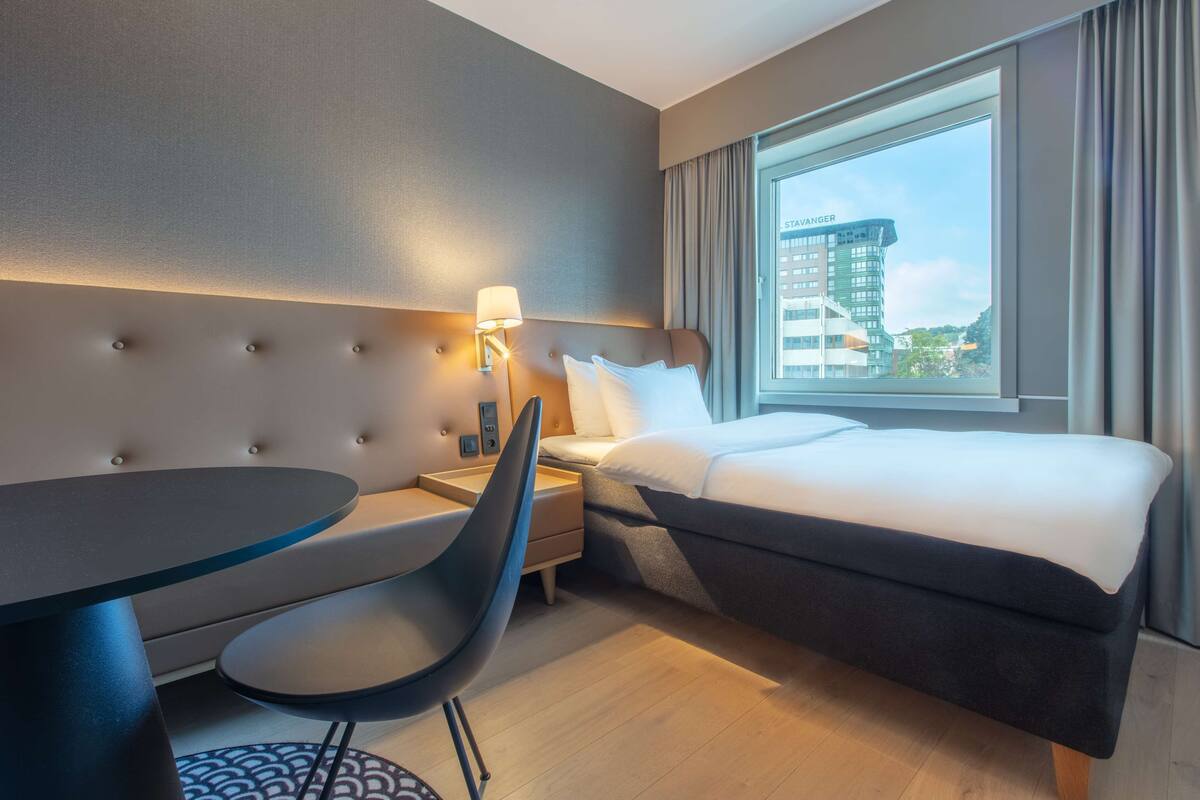 Moderni hotellihuone, jossa on sänky, työpöytä, tuoli ja ikkuna, josta on näkymä rakennukseen.