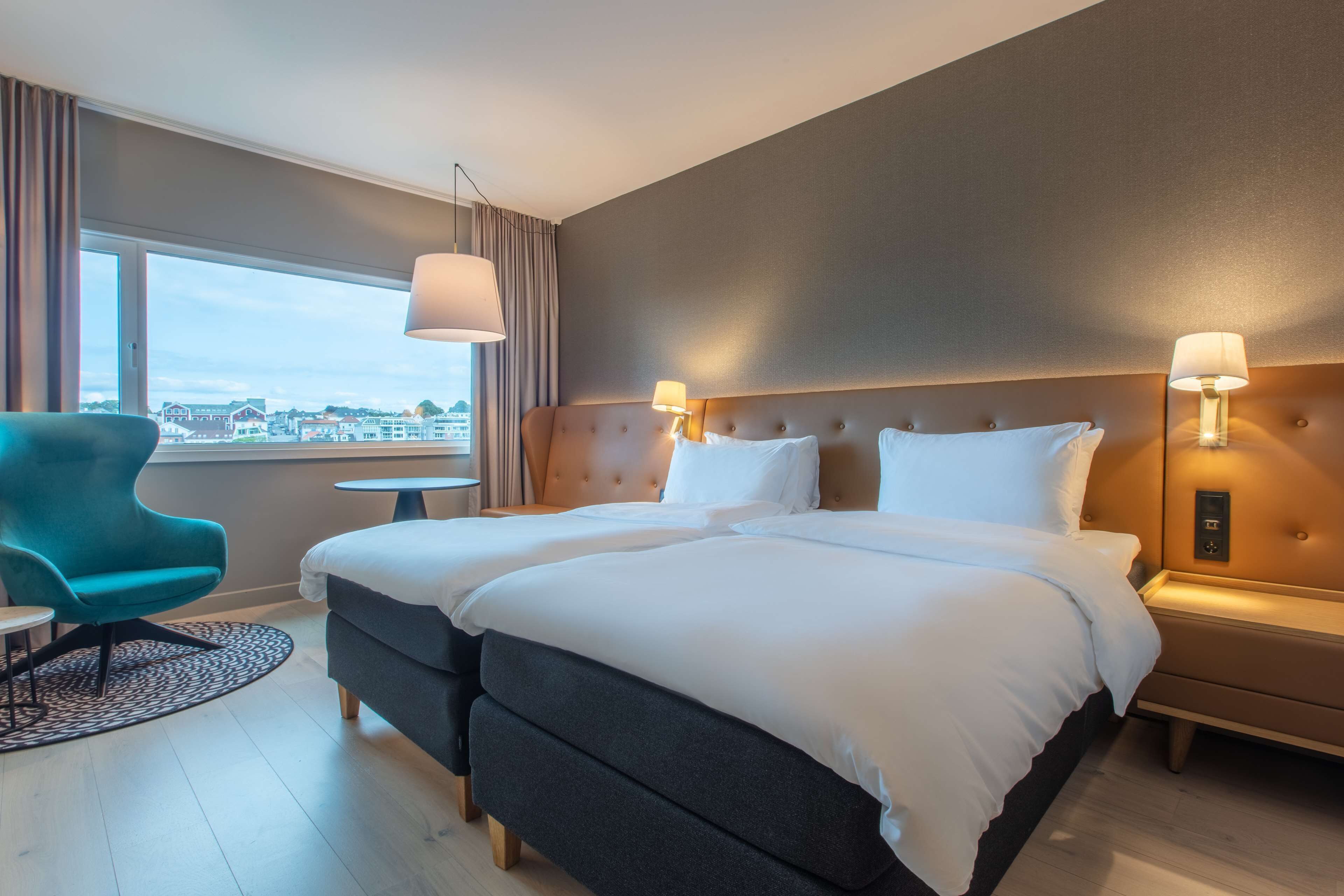 A hotel room with two beds, a teal armchair, a small table, and a view of the city.