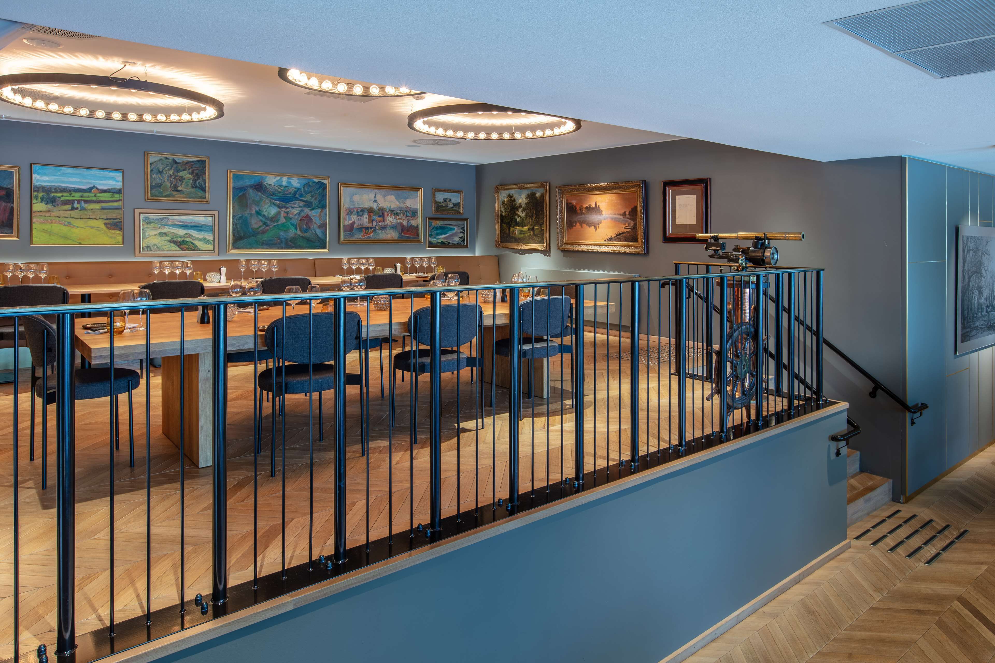A dining area with a long table, chairs, and a railing. There are framed pictures on the wall and a staircase with a railing.