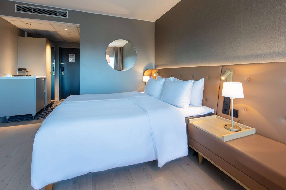 Moderni hotellihuone, jossa on suuri sänky, yöpöytä lampun kanssa, pyöreä peili seinällä ja pieni keittonurkkaus.