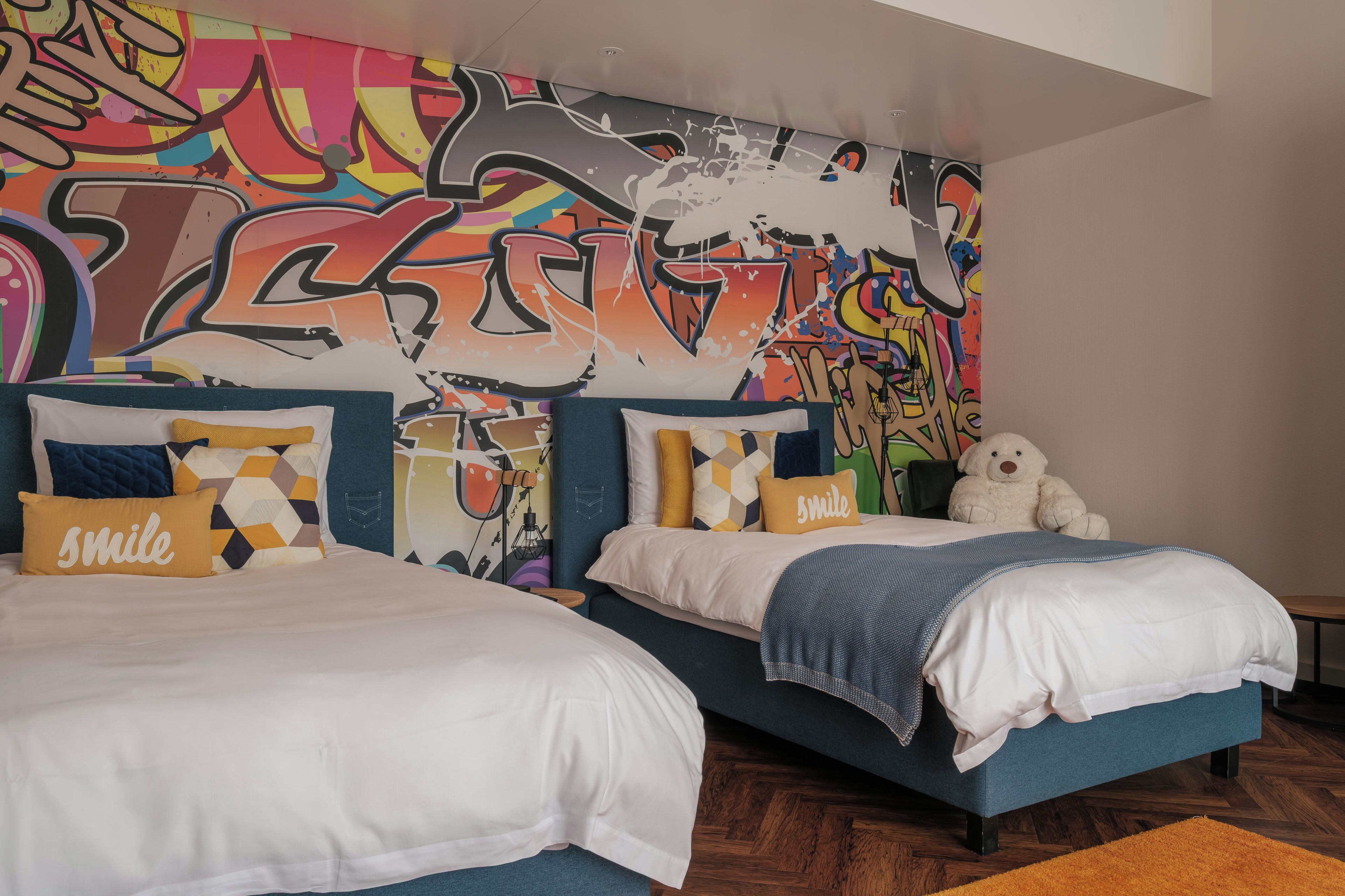 Quarto com duas camas, parede com grafite colorido e um urso de pelúcia.