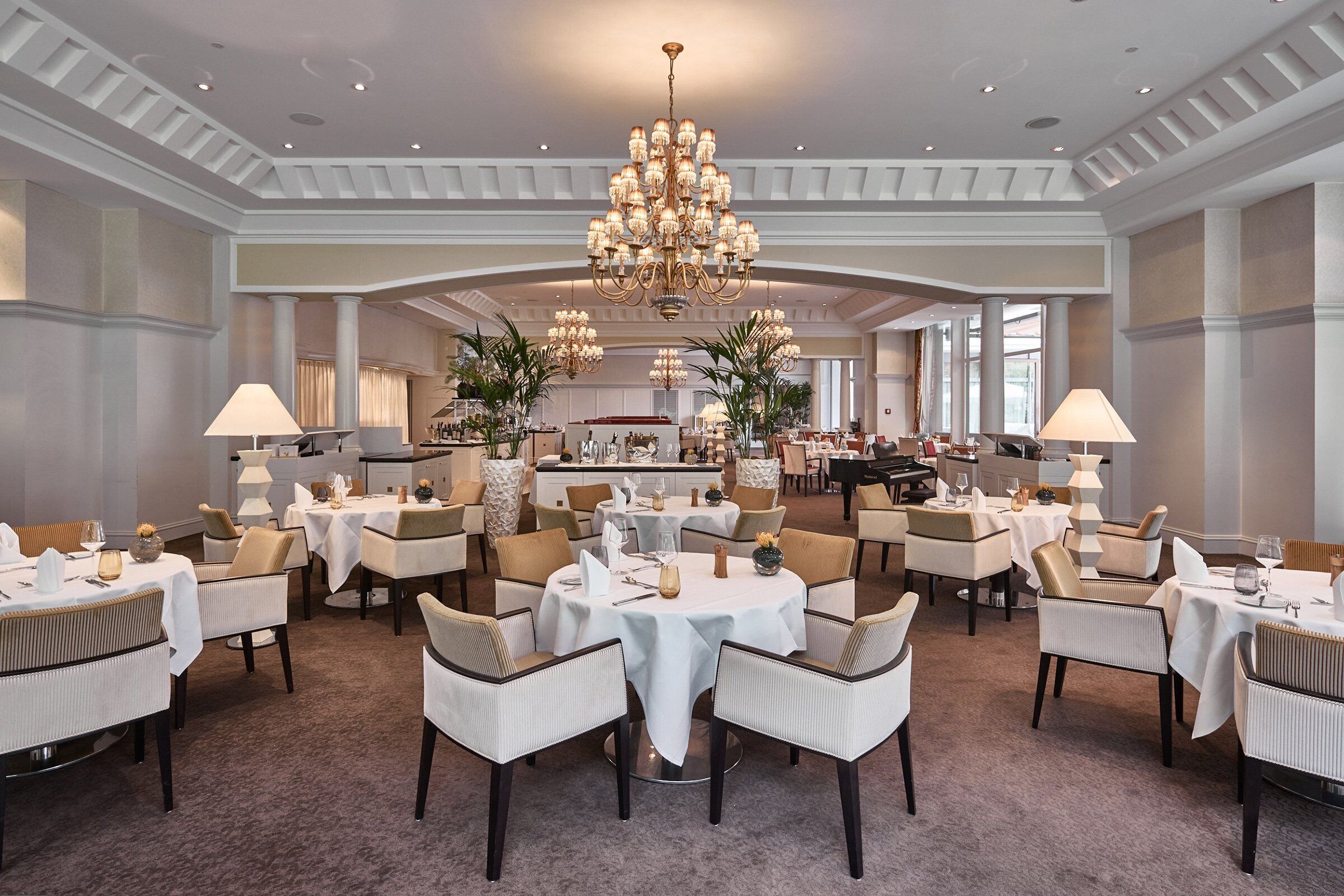 A spacious dining area with round tables and chairs, a large chandelier, and a view of the kitchen in the background.