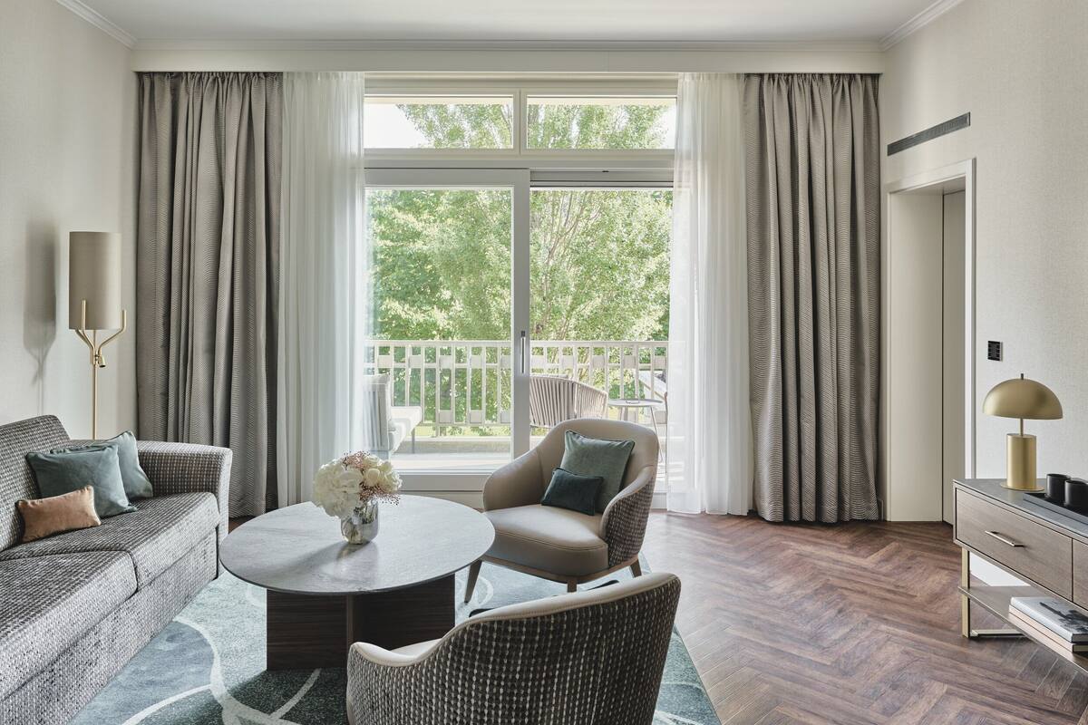 A modern living room with a sofa, armchair, and coffee table, a large window with curtains, and a view of the outdoors.