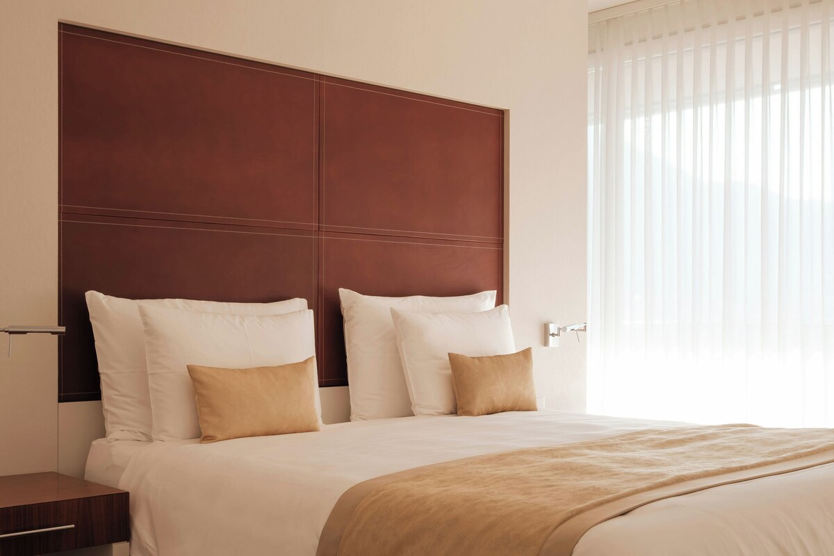 Habitación de hotel con una cama grande, cabecera de madera, mesita de noche y una ventana con cortinas traslúcidas.