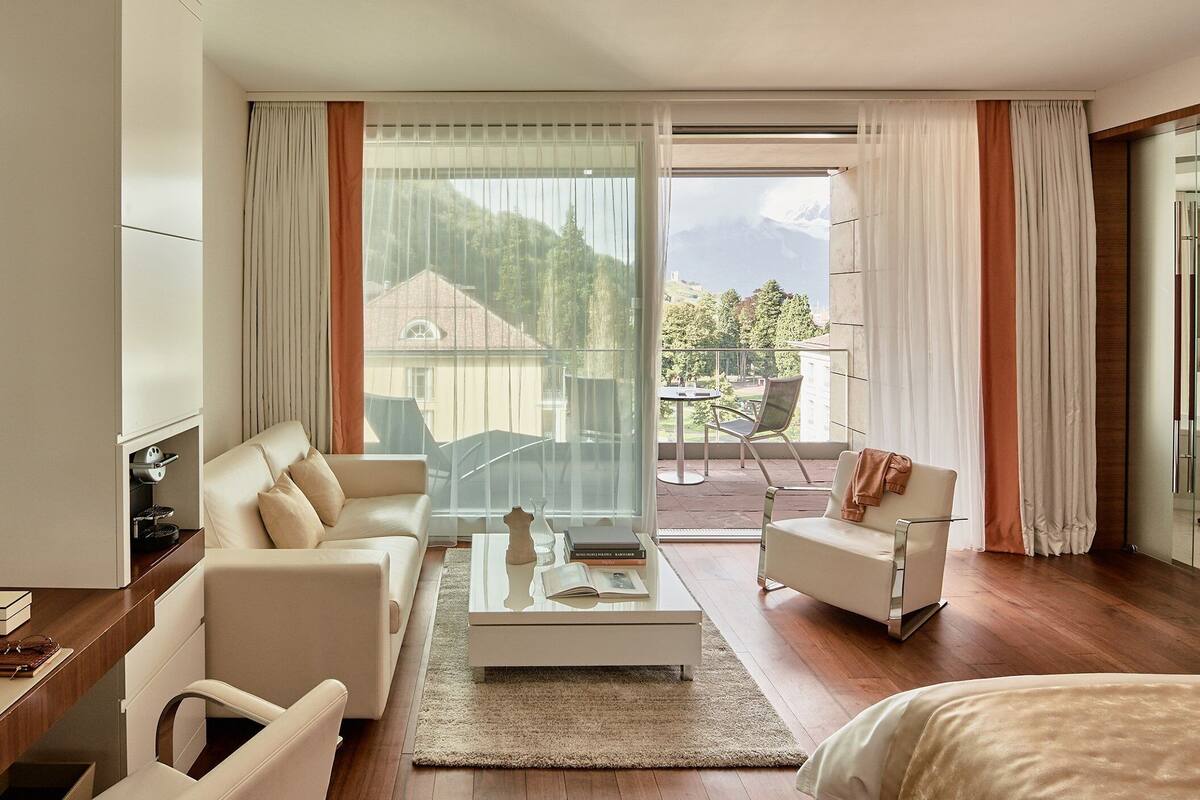 A modern hotel room with a sofa, armchair, and a view of a mountain landscape through large windows.