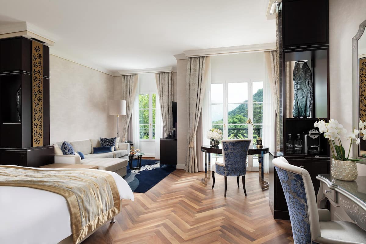 A hotel room with a large bed, a desk, a chair, and a view of greenery outside.