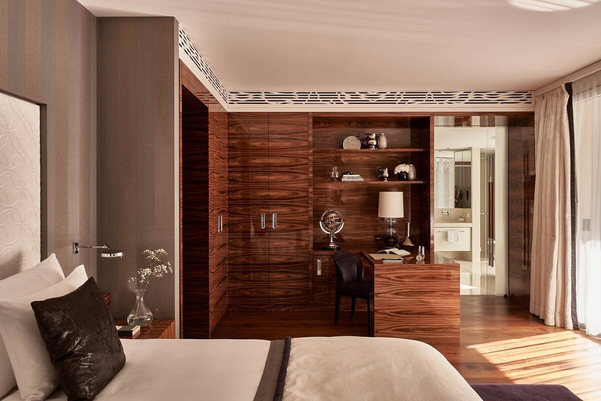A modern hotel room with a large bed, a wooden wardrobe, a desk with a lamp, and a vanity with a mirror.