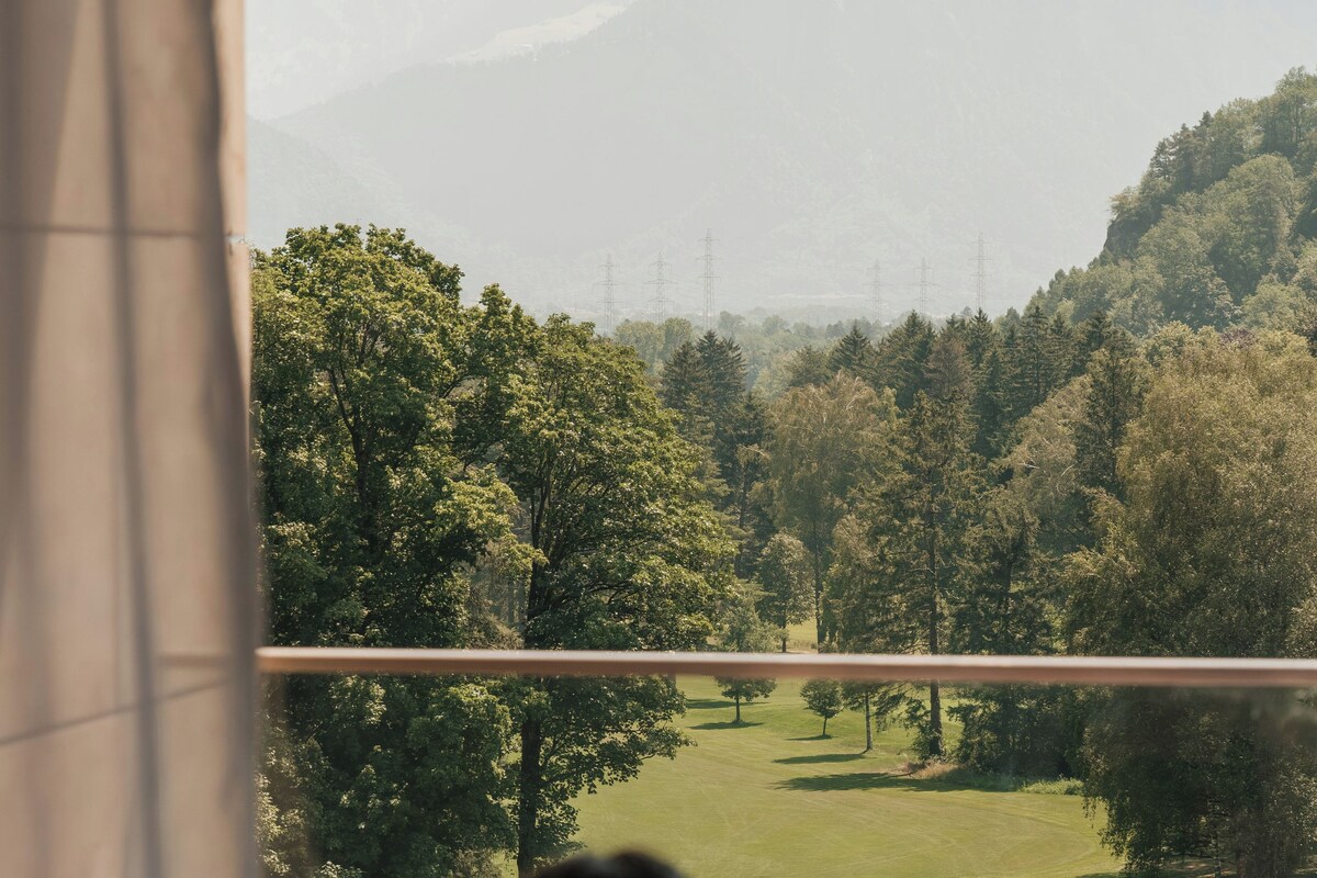 Una persona sentada junto a una ventana con vistas a un paisaje verde, árboles y un campo de golf.