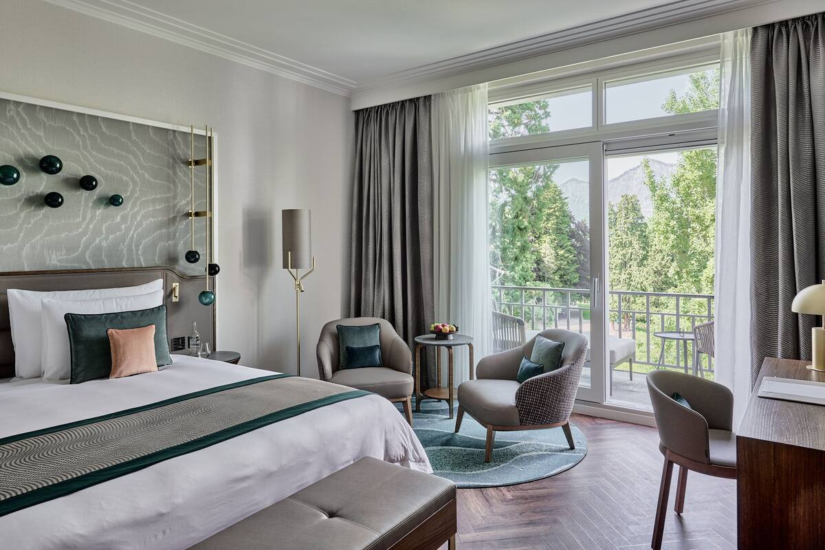 A hotel room with a large bed, two armchairs, a small table, and a view of the outdoors.