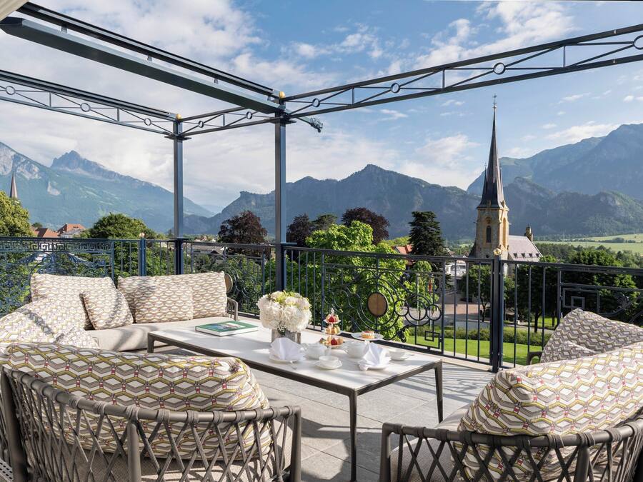 A terrace with wicker furniture, a table set with plates, cups, and a vase of flowers, overlooking a mountainous landscape with a church spire.