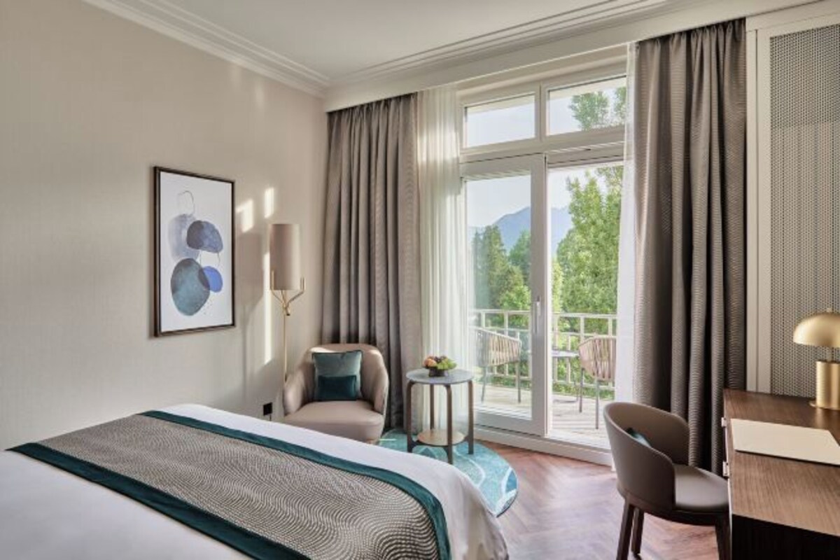 Habitación de hotel con cama, dos sillas, escritorio y balcón con vistas a árboles y montañas.