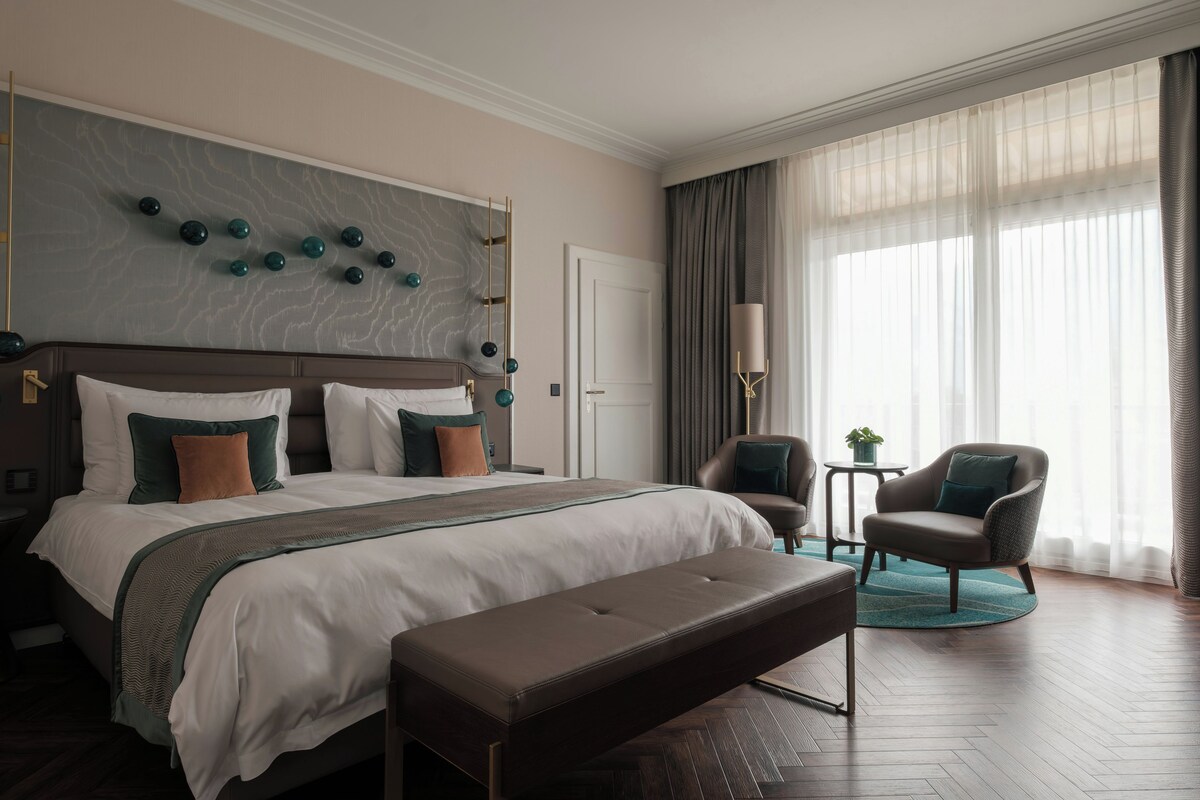 Habitación de hotel con una cama grande, un banco, dos sillones, una mesita y un ventanal con cortinas.