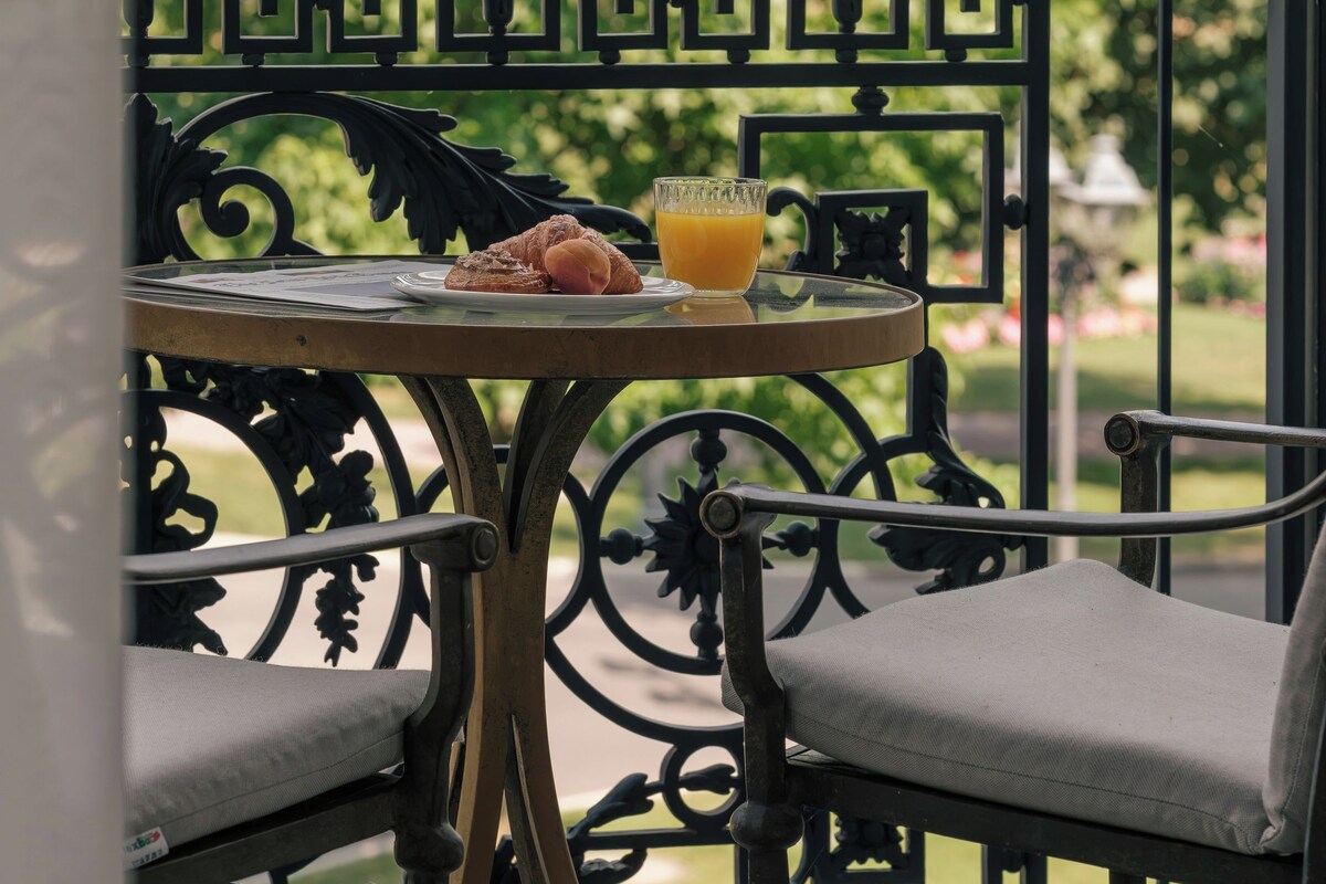 Una mesa con un vaso de jugo de naranja y un croissant, en un balcón con barandilla metálica y vistas a la vegetación.