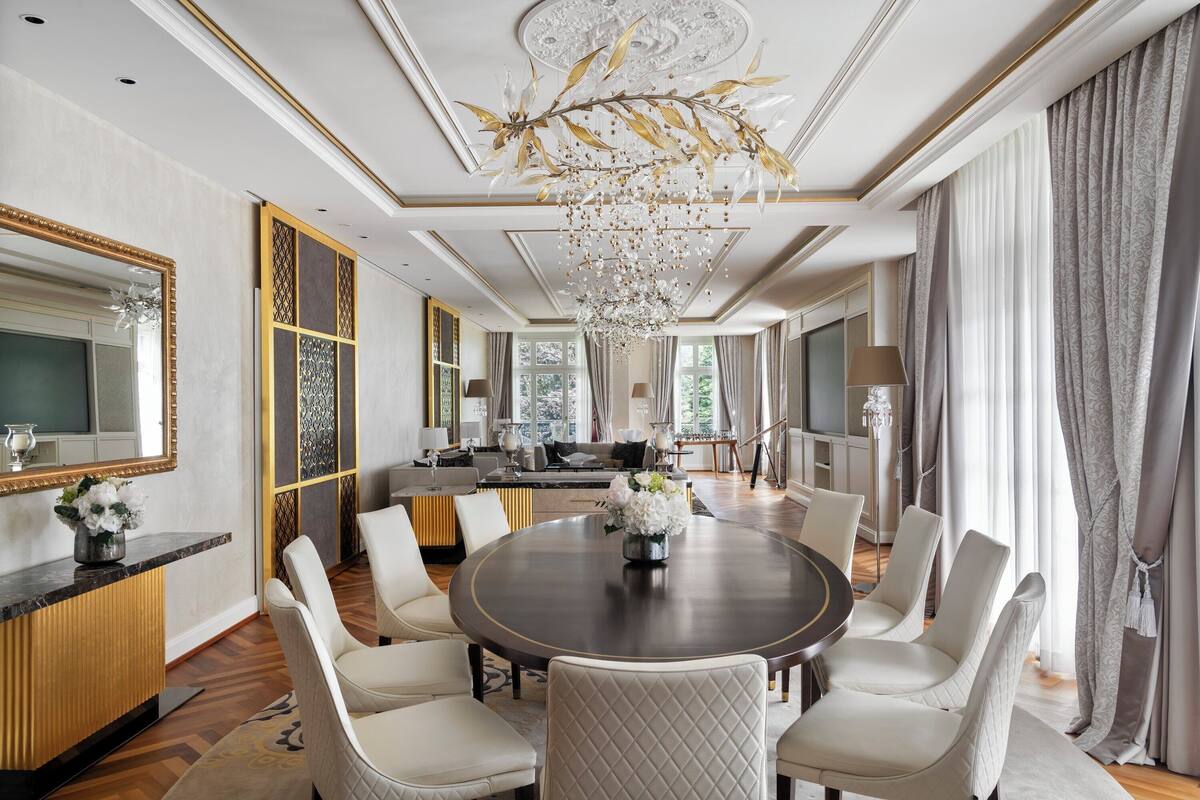 A luxurious dining room with a round table, white upholstered chairs, and a chandelier.