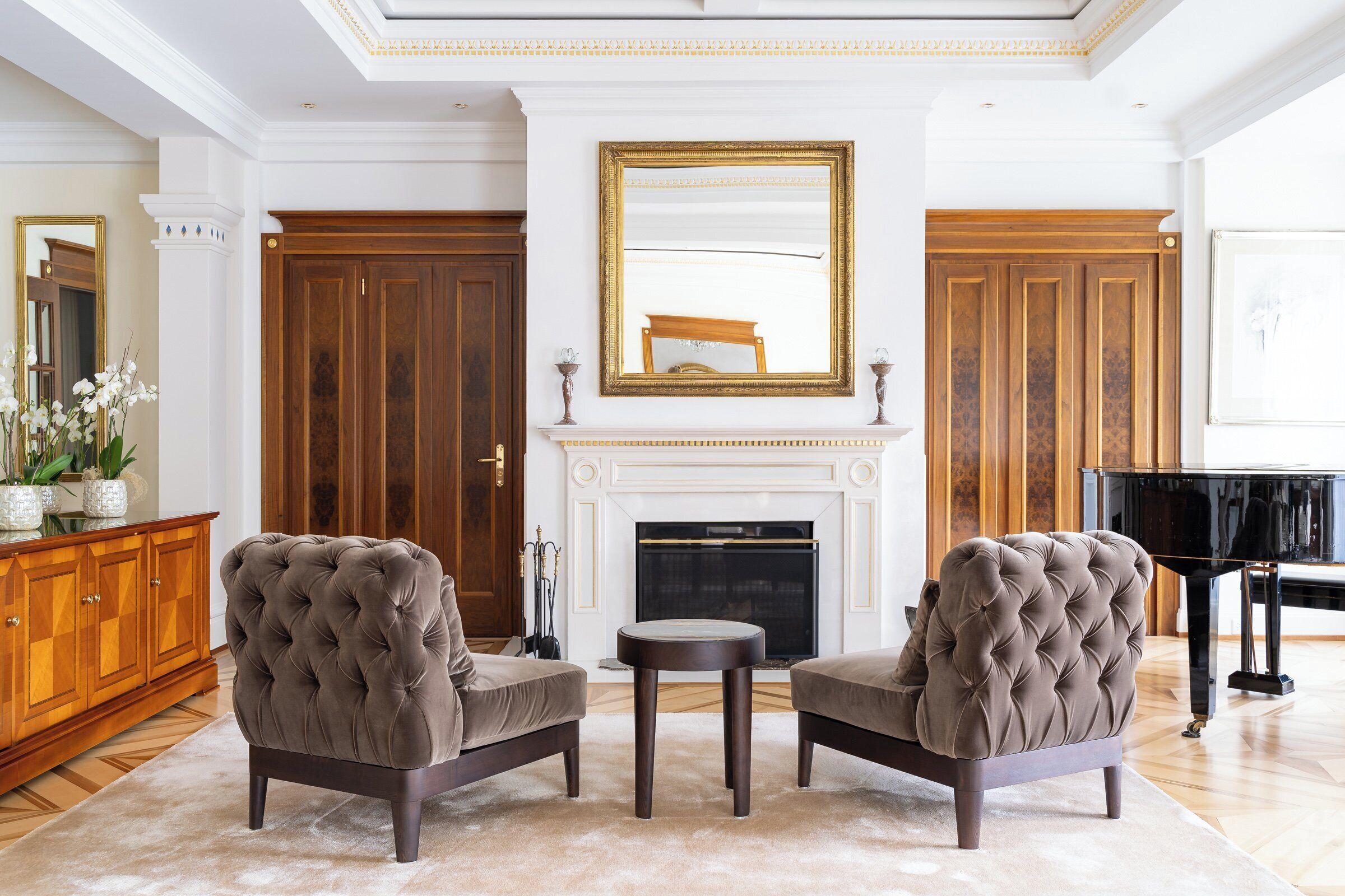 A living room with two tufted armchairs, a fireplace, a grand piano, and a large mirror.