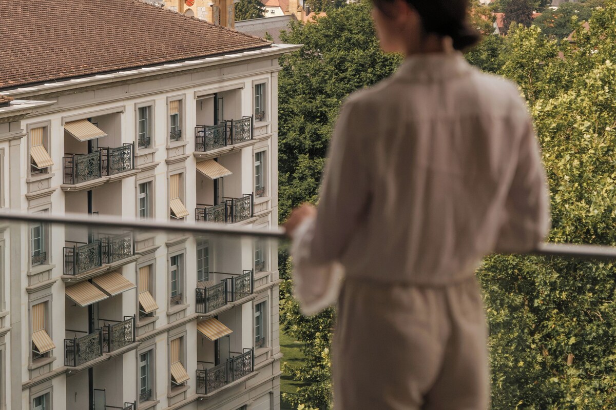 Una persona observa un edificio con una torre, rodeado de vegetación y montañas.