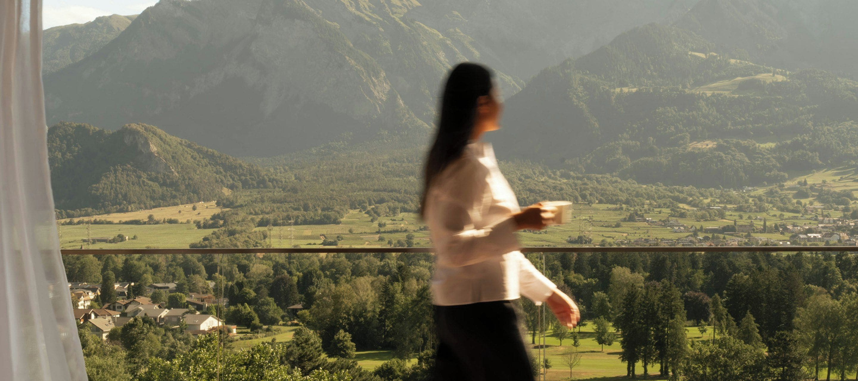 Una persona sostiene una taza, con vistas a un valle con casas y montañas al fondo.