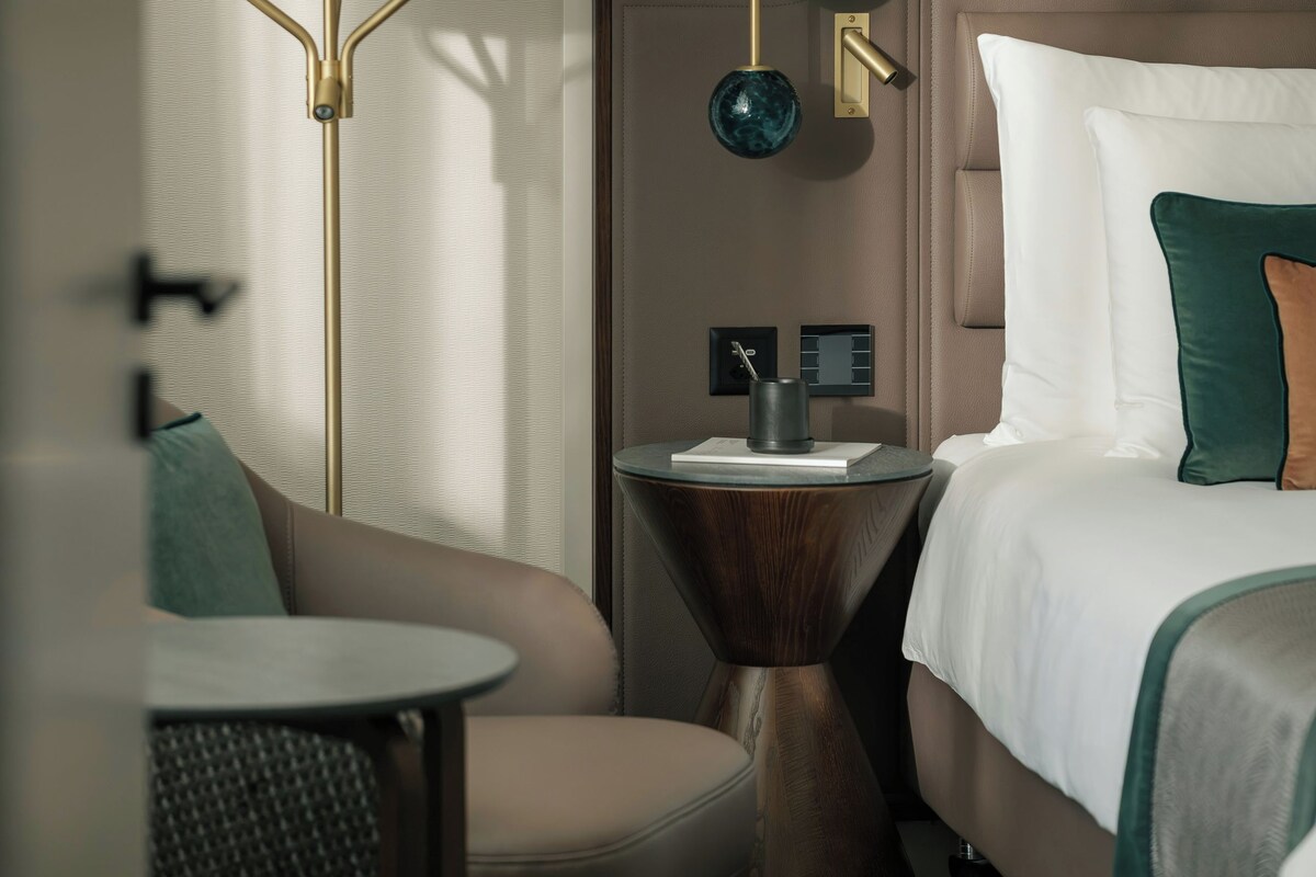 Habitación de hotel con cama, una mesa redonda, una silla, lámpara y apliques de pared.