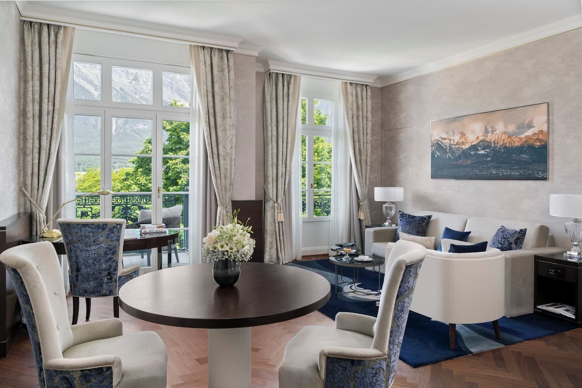 A modern living room with a round table, two armchairs, a sofa, and a large window with curtains.