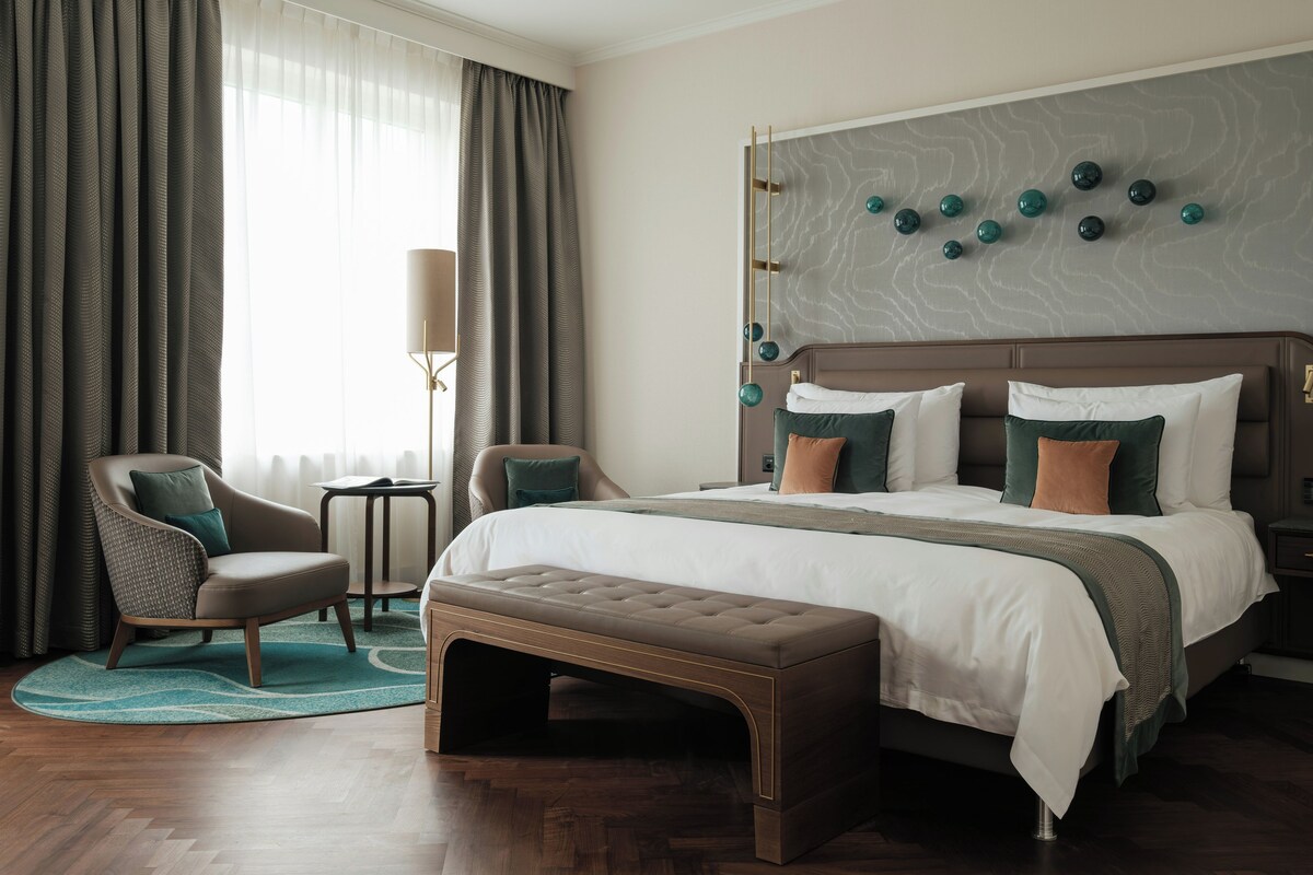 Habitación de hotel con una cama grande, una zona de estar con una silla y una mesita, y un adorno en la pared sobre la cama.