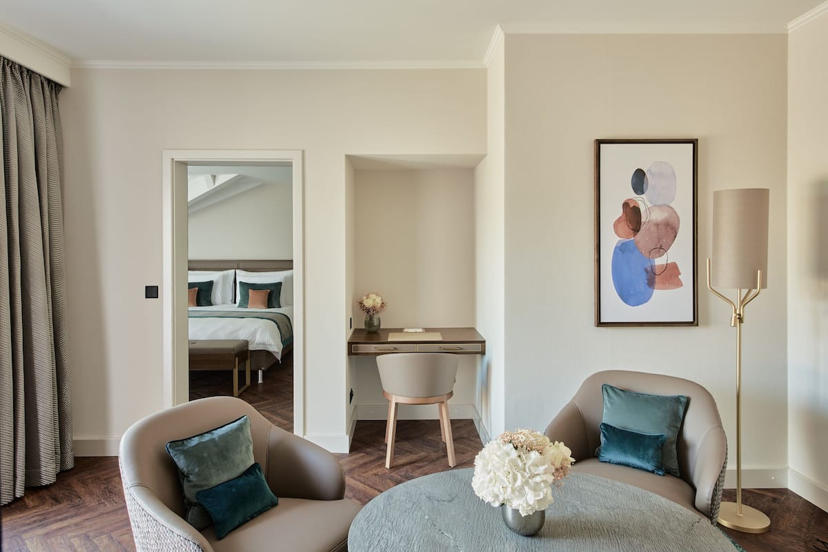 A hotel room with a bed, a desk, two armchairs, a round table with flowers, a lamp, and a painting on the wall.