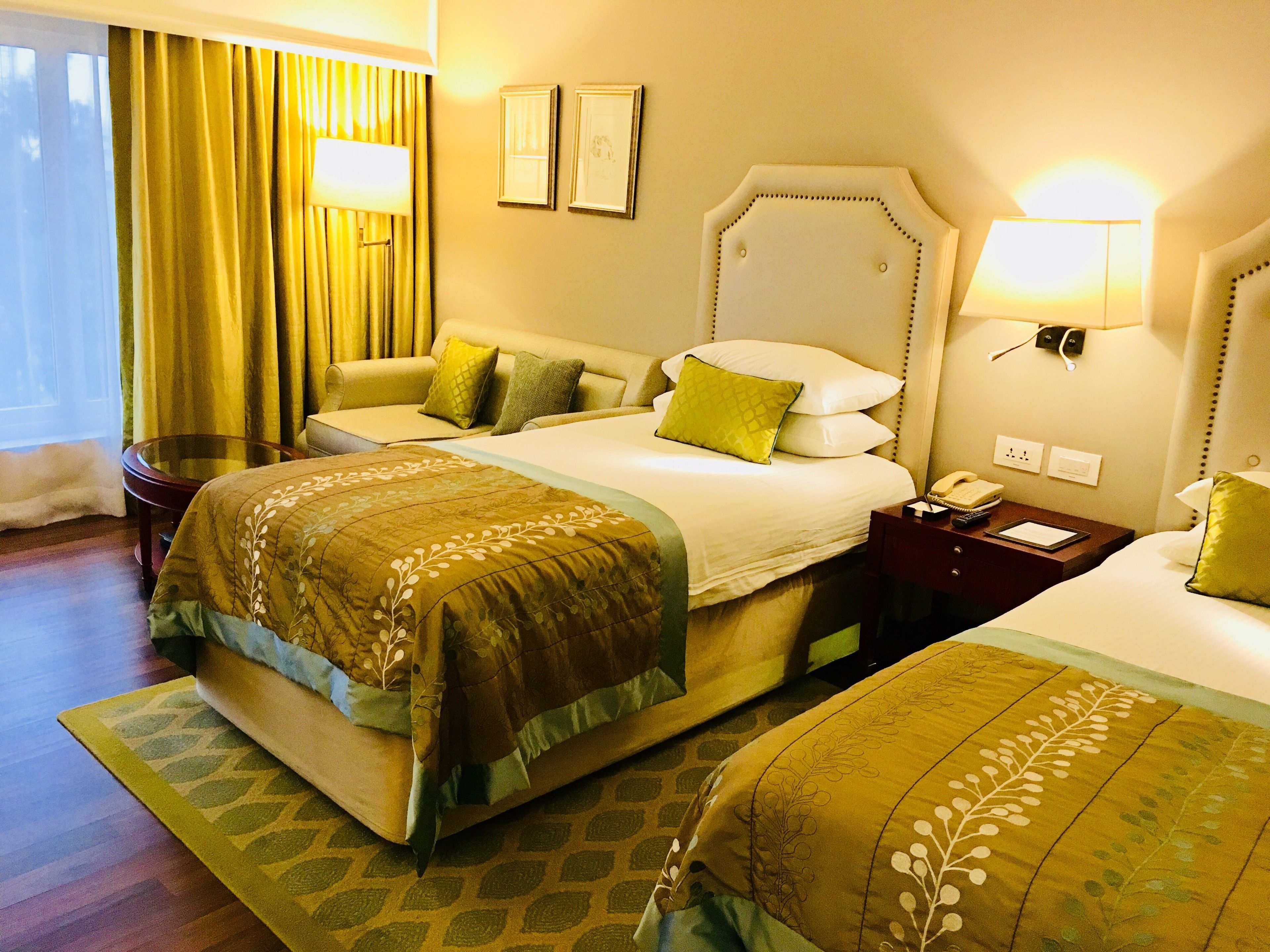 A hotel room with a large bed, a smaller bed, a bedside table with a lamp, a nightstand with a phone, and a window with curtains.