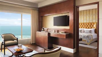Habitación de hotel con un ventanal grande con vista al océano, un televisor de pantalla plana montado en la pared, una cama con cabecera capitonada y una zona de comedor con mesa y sillas.