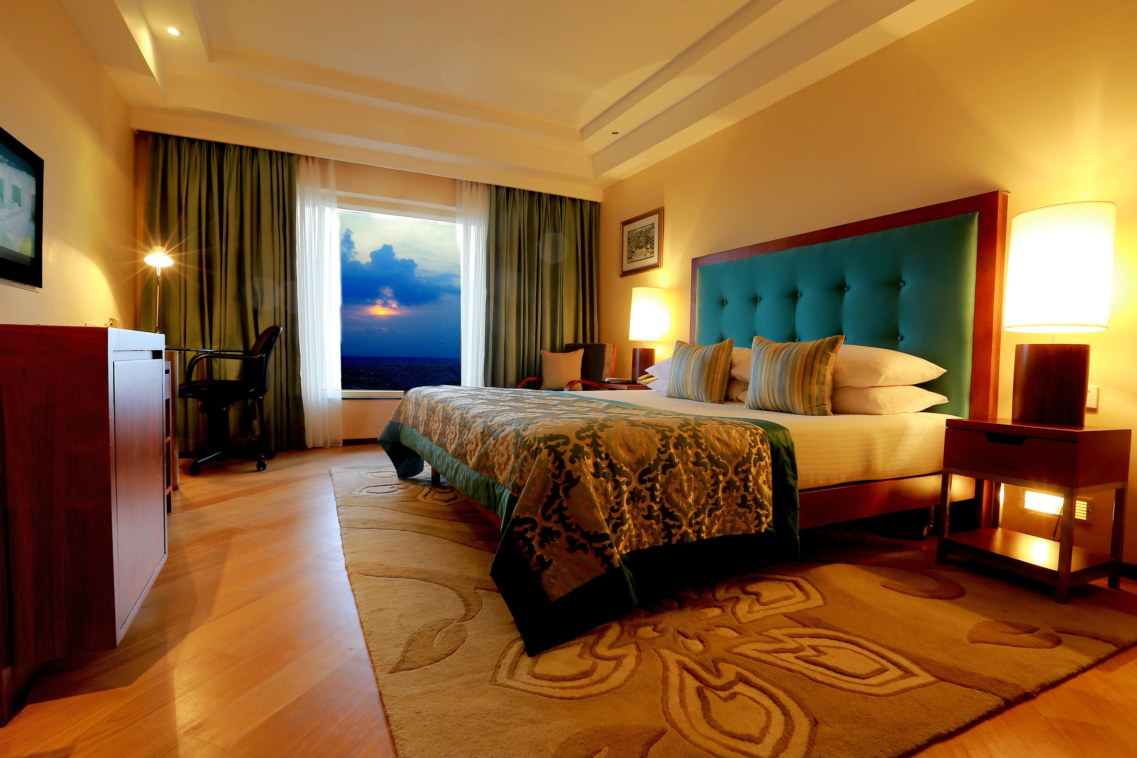 A hotel room with a large bed, a desk, a chair, and a view of the sunset through the window.