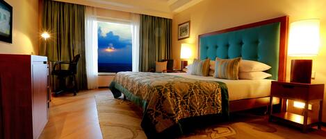 A hotel room with a large bed, a desk, a chair, and a view of the sunset through the window.
