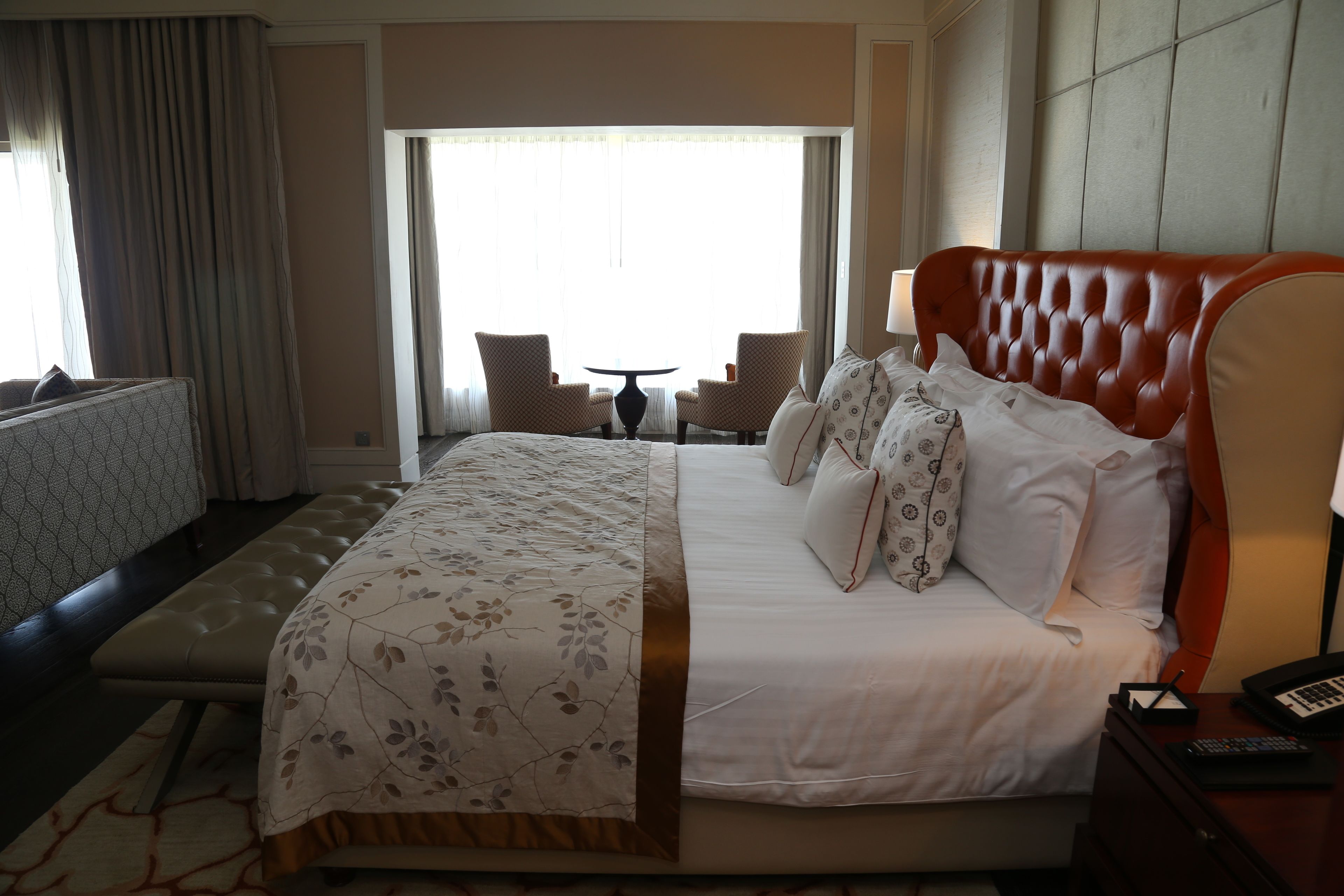 A hotel room with a large bed, a headboard, a bedside table with a lamp, a small table with chairs, and a window with curtains.