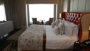 A hotel room with a large bed, a headboard, a bedside table with a lamp, a small table with chairs, and a window with curtains.