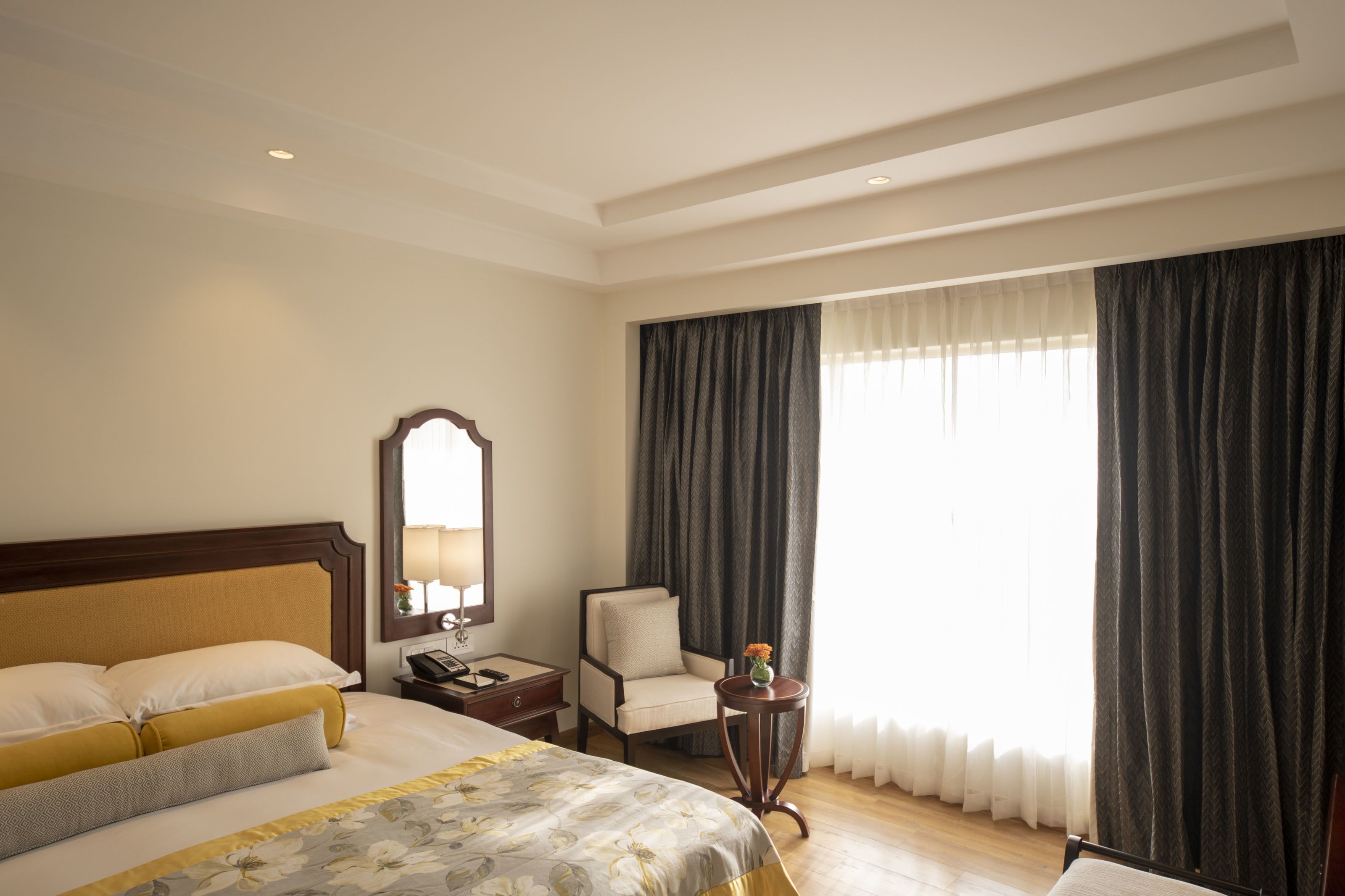 A hotel room with a large bed, a chair, a small table, a nightstand, and a window with curtains.