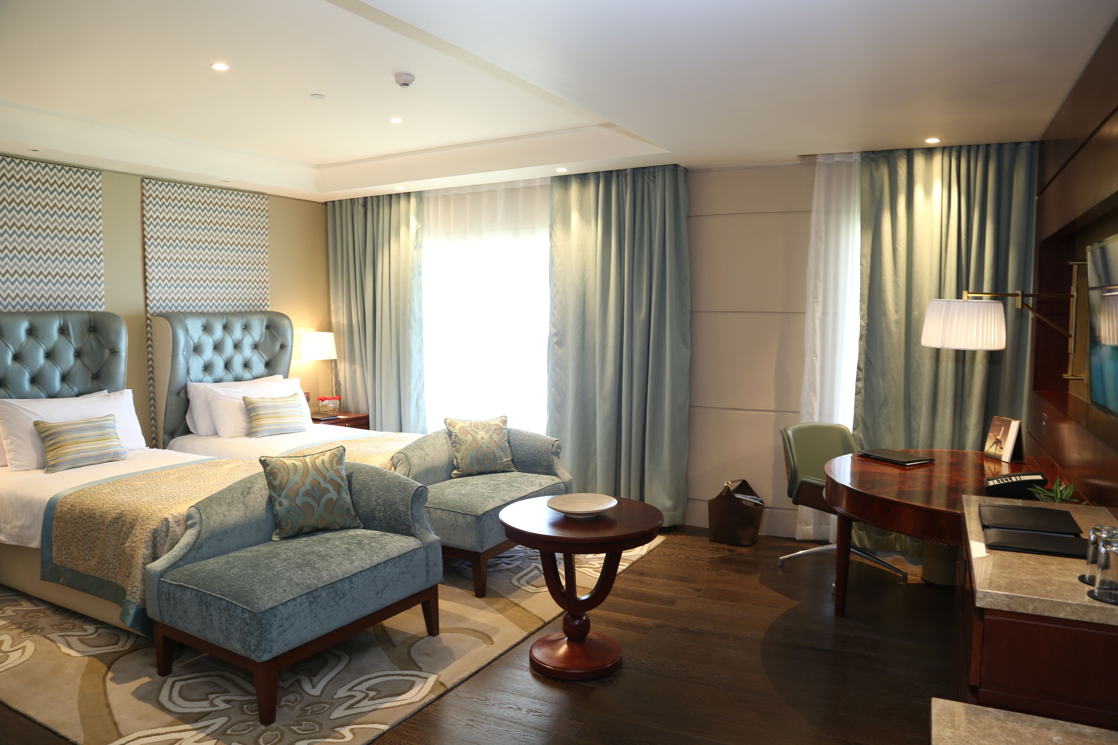 A hotel room with a large bed, two armchairs, a small round table, a desk with a chair, and a large window with curtains.