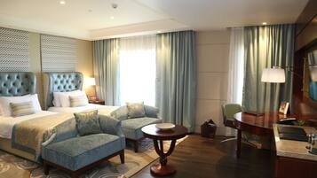 A hotel room with a large bed, two armchairs, a small round table, a desk with a chair, and a large window with curtains.