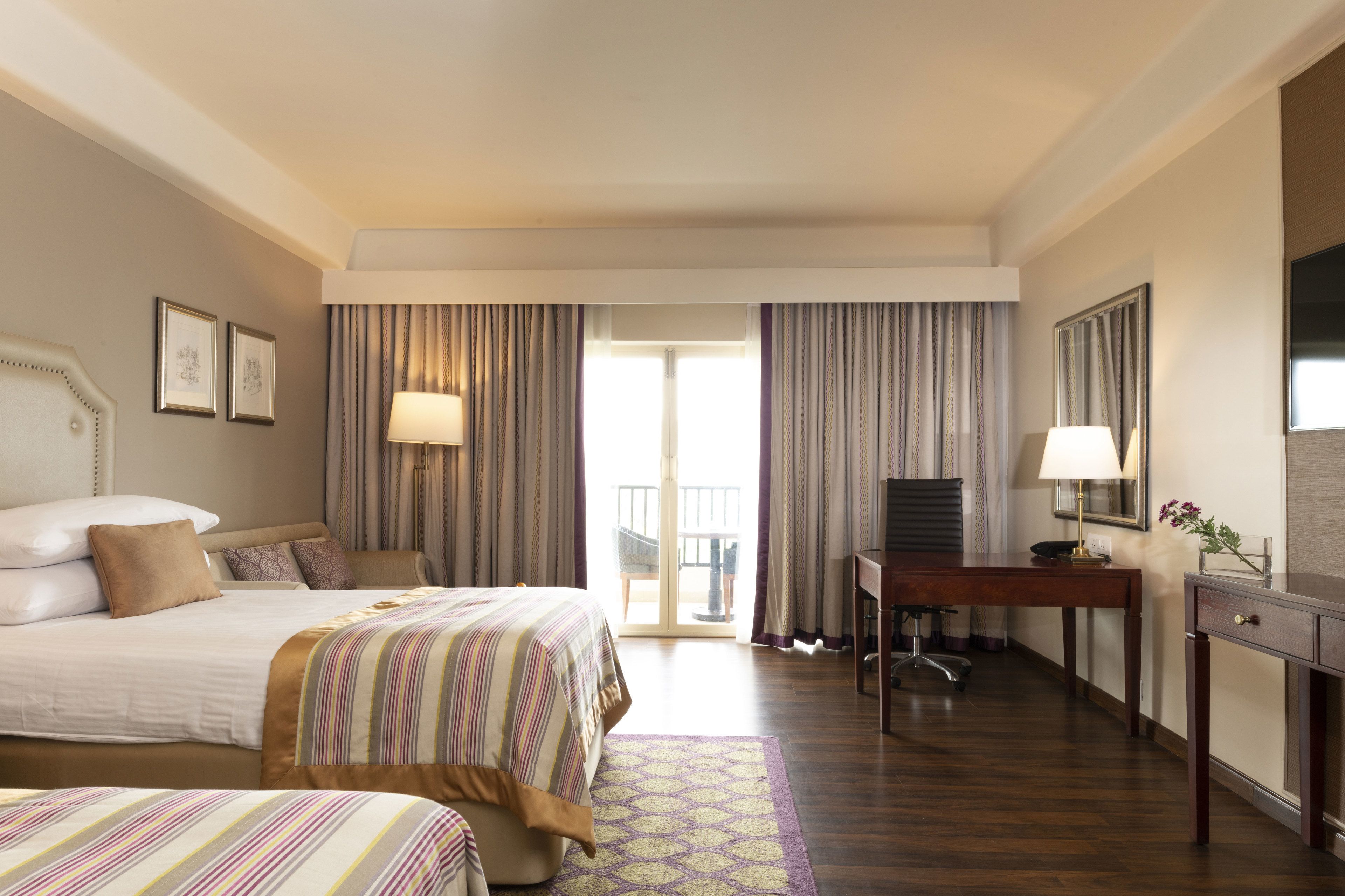 Habitación de hotel con cama, escritorio y silla. Cuenta con una ventana con cortinas, una puerta que da a un balcón y un televisor montado en la pared.