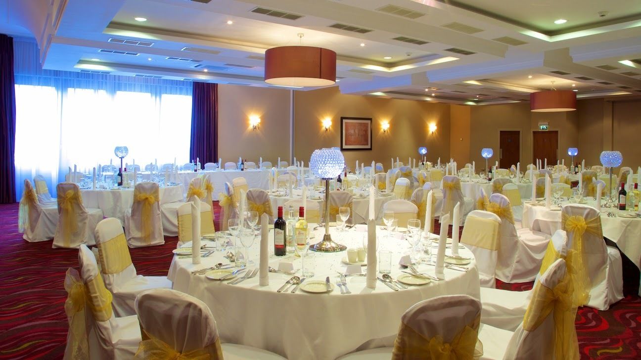 A banquet hall with round tables set for a formal event, featuring white tablecloths, gold sashes, and candle centerpieces.