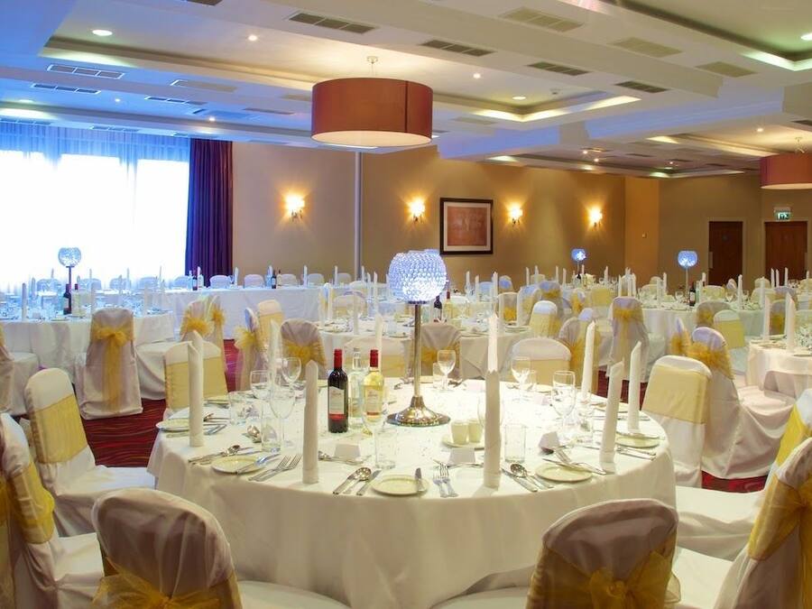Een balzaal met ronde tafels gedekt voor een formeel evenement, met witte tafelkleden, gouden linten en kaarsen als middelpunt.