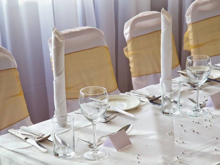 Una mesa dispuesta para un evento formal, con manteles blancos, fajas doradas y copas de cristal.